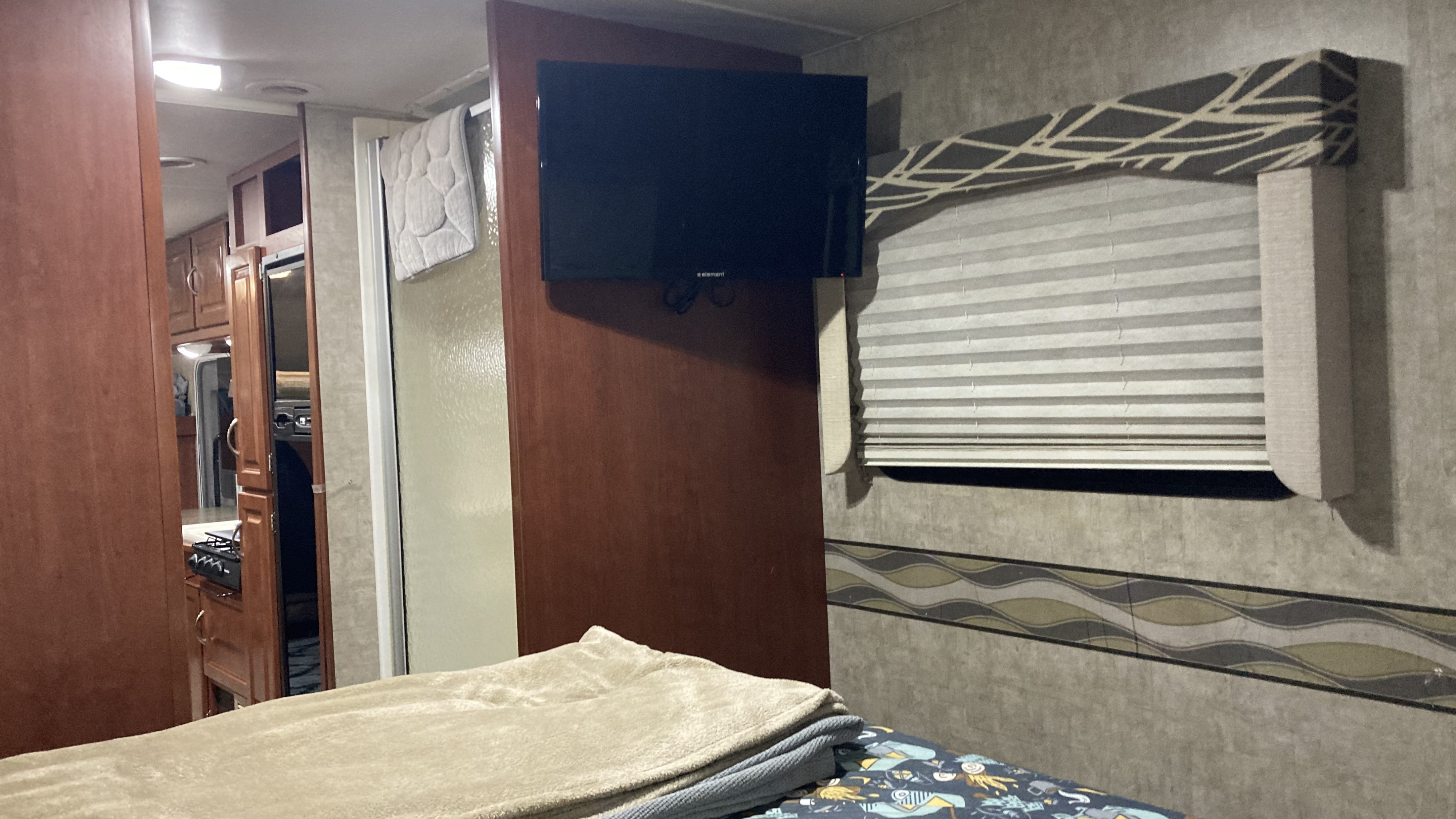 Cozy RV rental interior: bed with blankets, wall-mounted TV, kitchen glimpse, and window with blinds.. Winnebago Minnie Winnie 2014