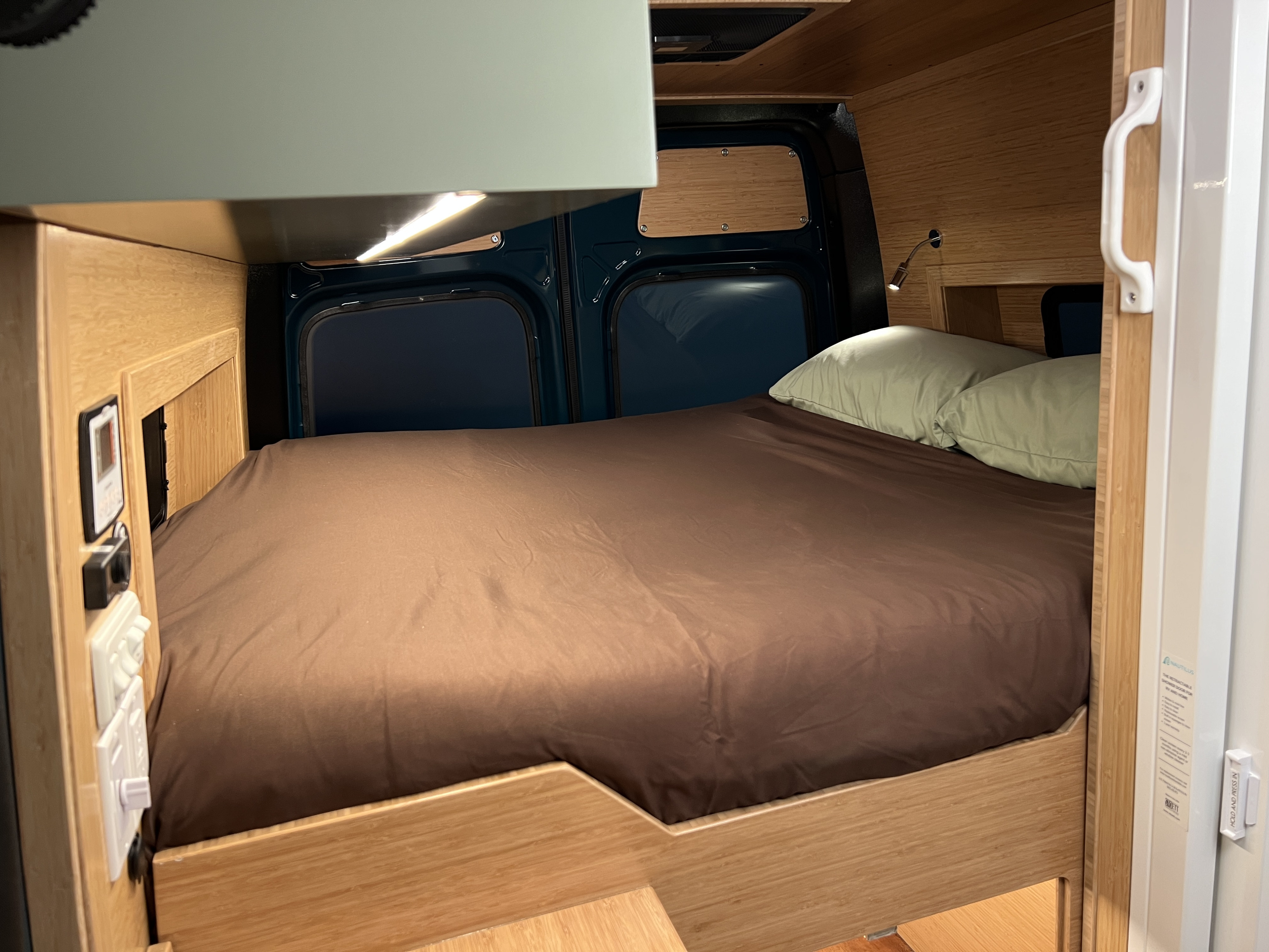 Inside a modern rv rental campervan: a comfy bed with wood paneling, overhead lighting, and controls for a perfect road trip.. Mercedes-Benz Sprinter 2026