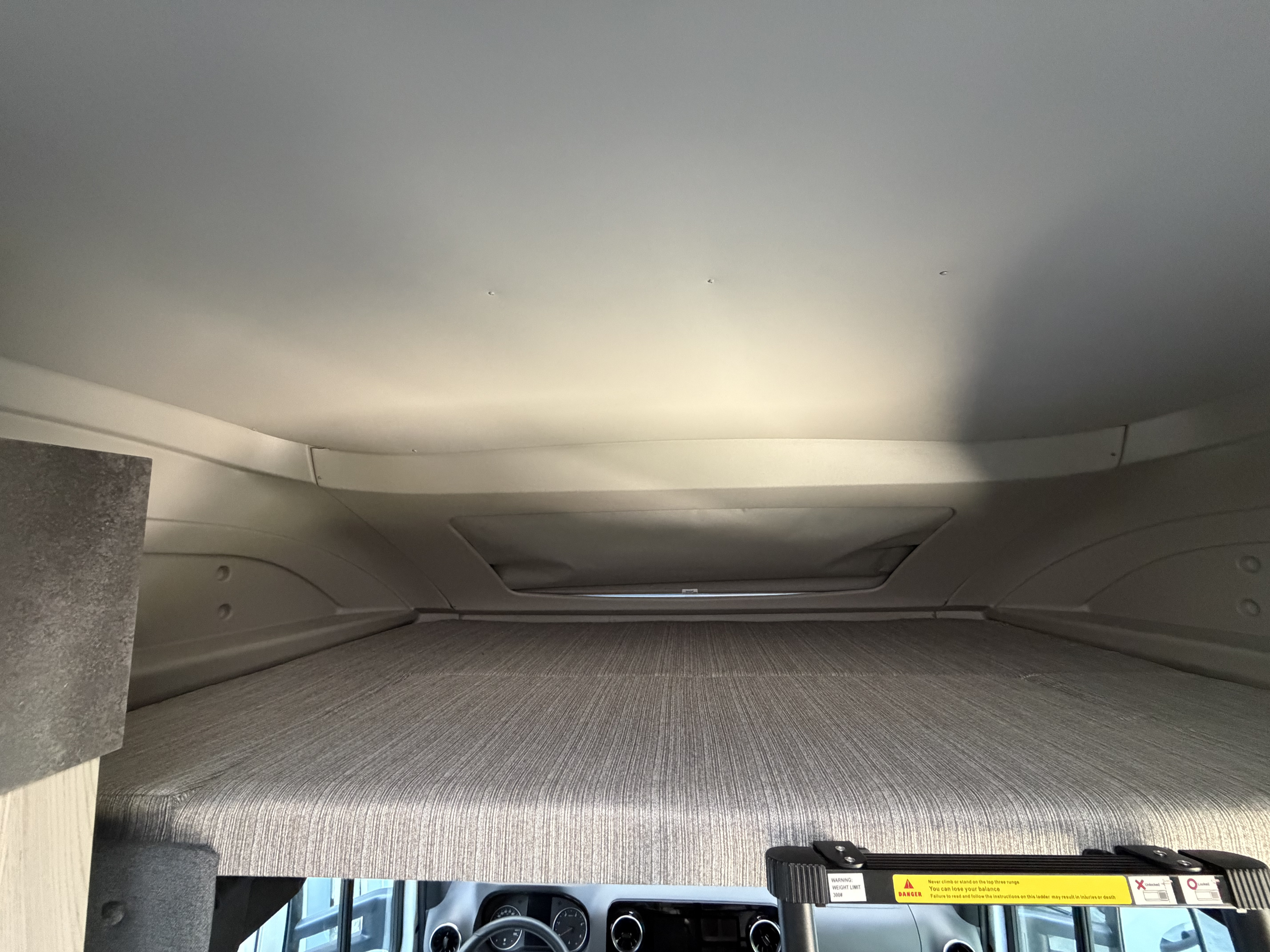 Inside an RV rental: the cab-over bunk with a textured mattress, curved ceiling, and a ladder with a 300# weight limit warning.. Coachmen Prism 2026