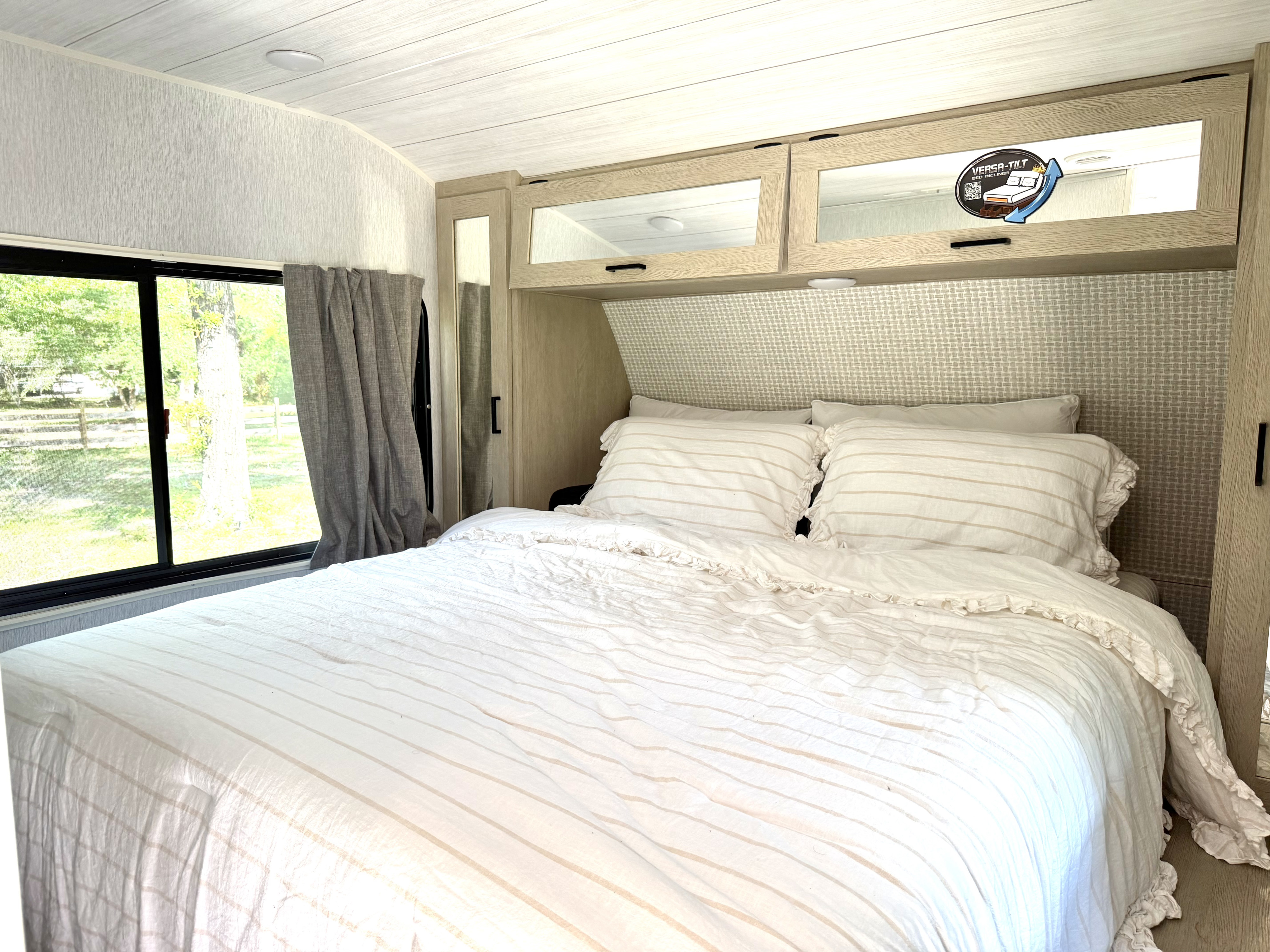 This RV rental bedroom features a comfy bed with striped bedding, light wood overhead storage, and a large window showcasing nature.. Forest River Salem Platinum 2024