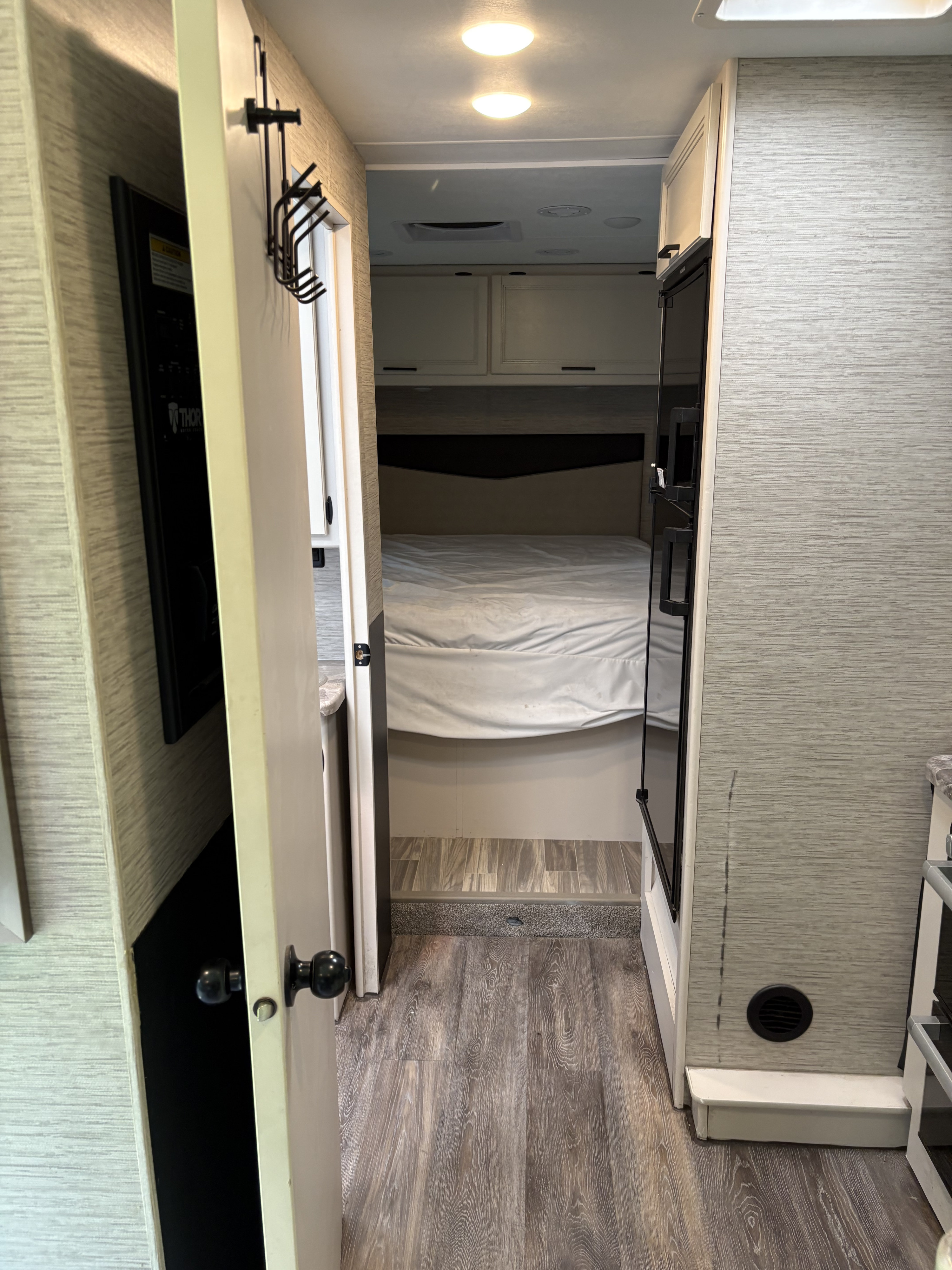 RV rental interior: Hallway view into a compact bedroom with bed & cabinets. Features wood-look floor, coat rack & refrigerator.. Thor Four Winds 2022