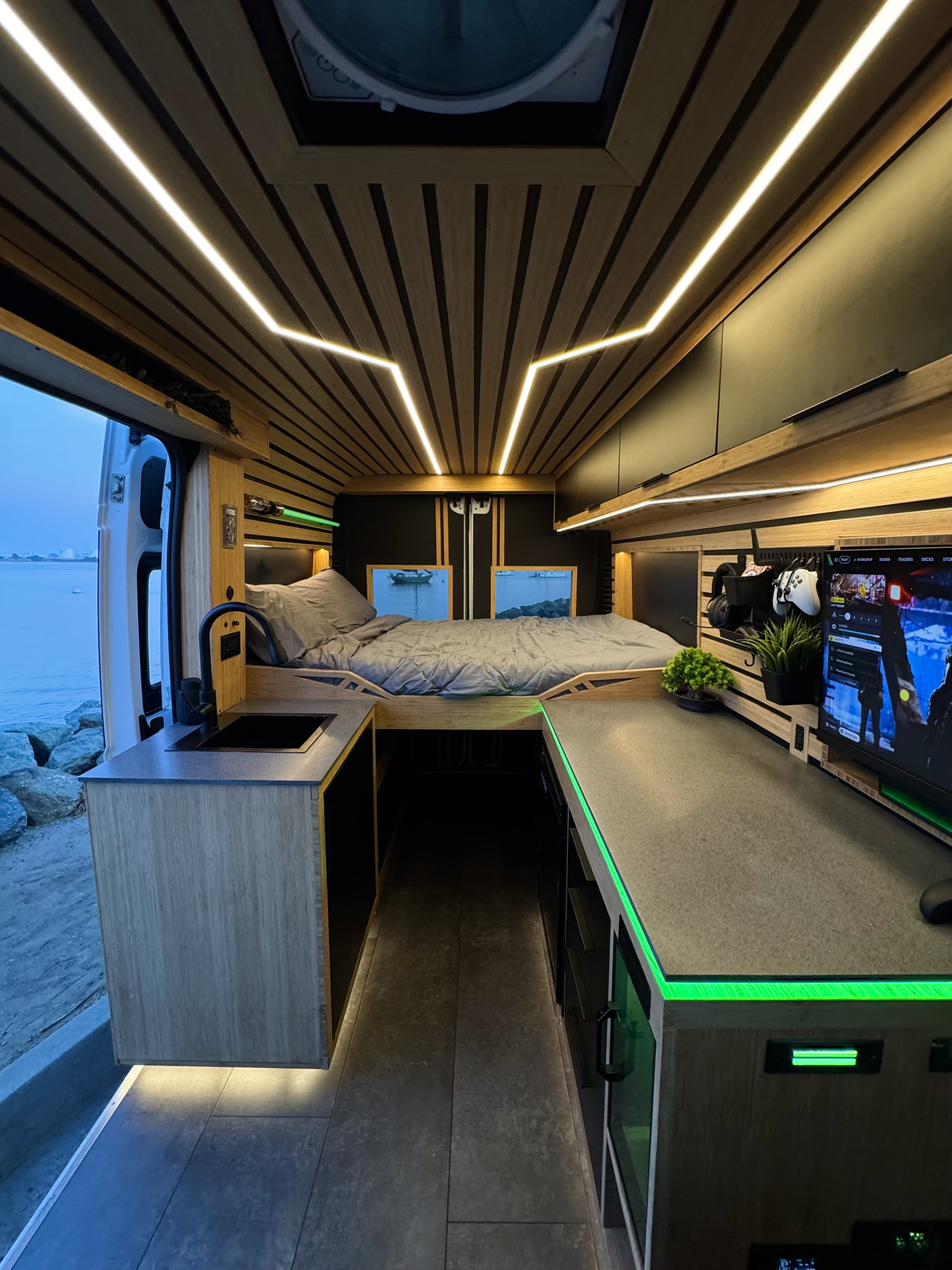 Modern custom RV rental interior with wooden ceiling, LED lighting, cozy bed, kitchen sink, and a cool gaming setup by the water.. Dodge Ram 1500 2019