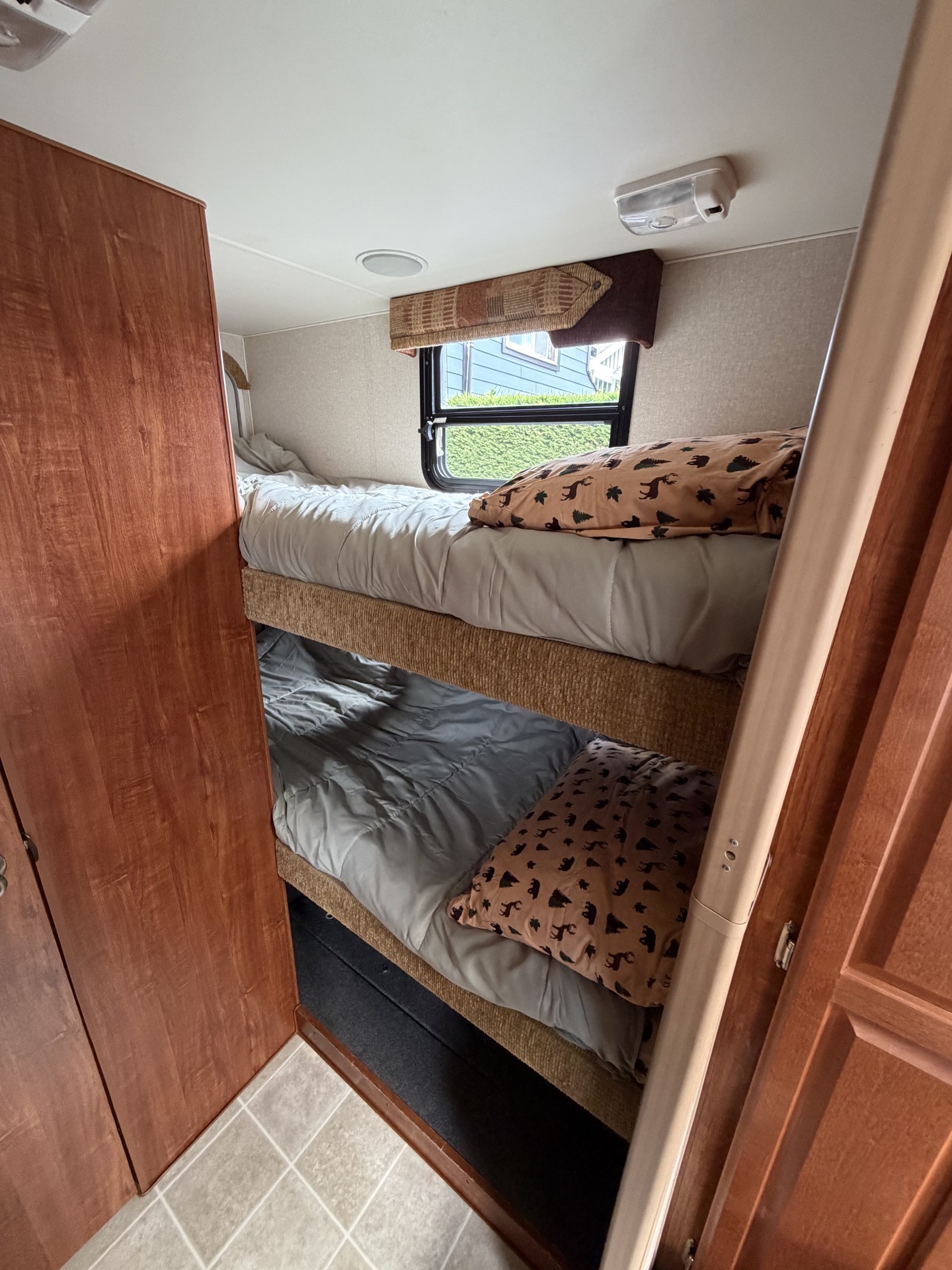An RV rental interior featuring cozy bunk beds with light-colored bedding, patterned pillows, and a window displaying green foliage.. Komfort Trailblazer 2005