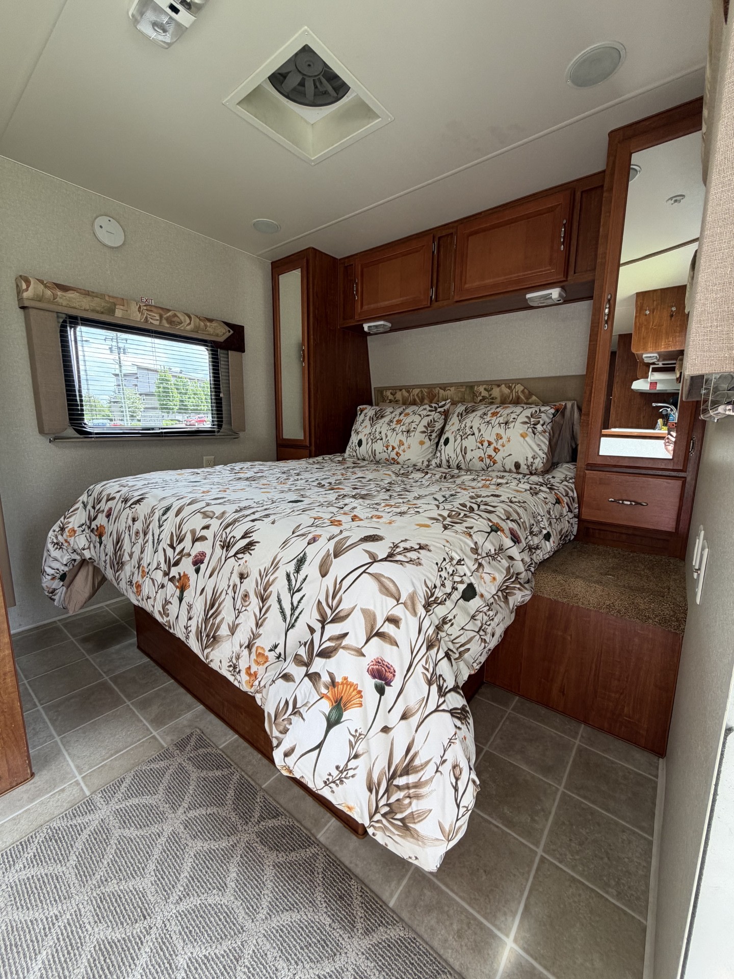 RV rental bedroom. Features a bed with white floral duvet, wood cabinets, window with blinds, mirror, and ceiling vent.. Komfort Trailblazer 2005