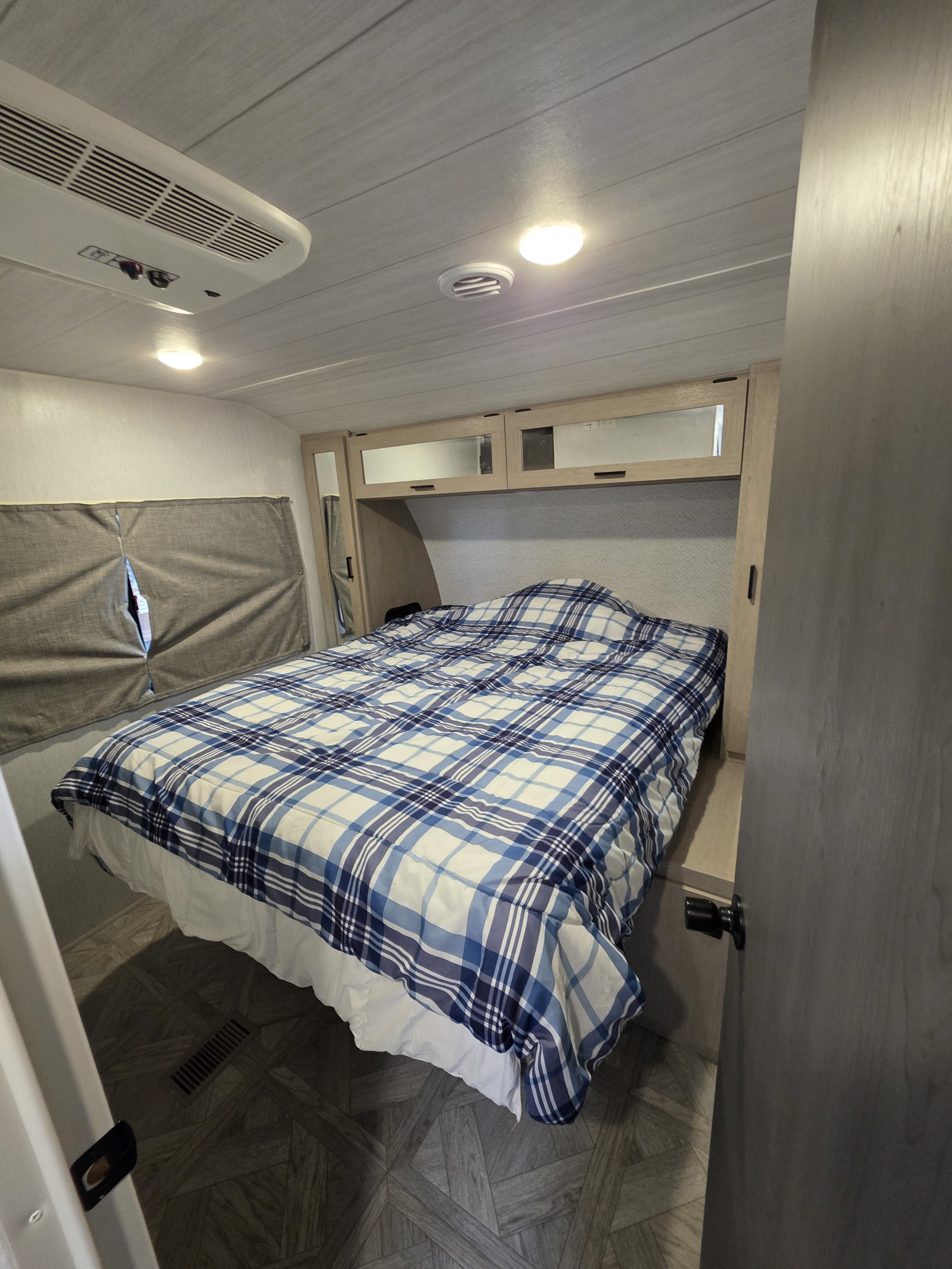 Inside a cozy RV rental bedroom: blue plaid bed, mirrored cabinets, AC, and shaded window for comfort.. Forest River Wildwood Platinum 2024
