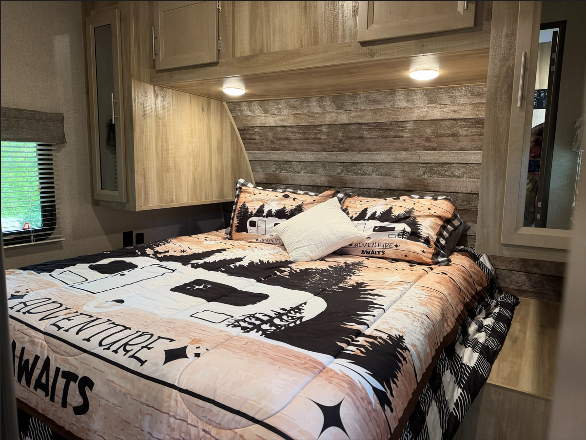 This cozy RV rental bedroom features rustic decor, a bed with an "Adventure Awaits" comforter, and ample overhead storage.. Coachmen Catalina Legacy 2022