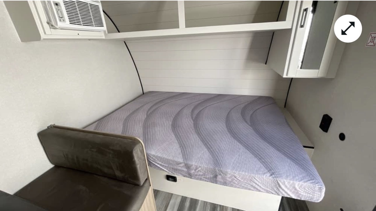 This rv rental shows a compact interior: bed with patterned sheet, window AC unit, overhead storage, and bench.. Jayco Jayco 2026