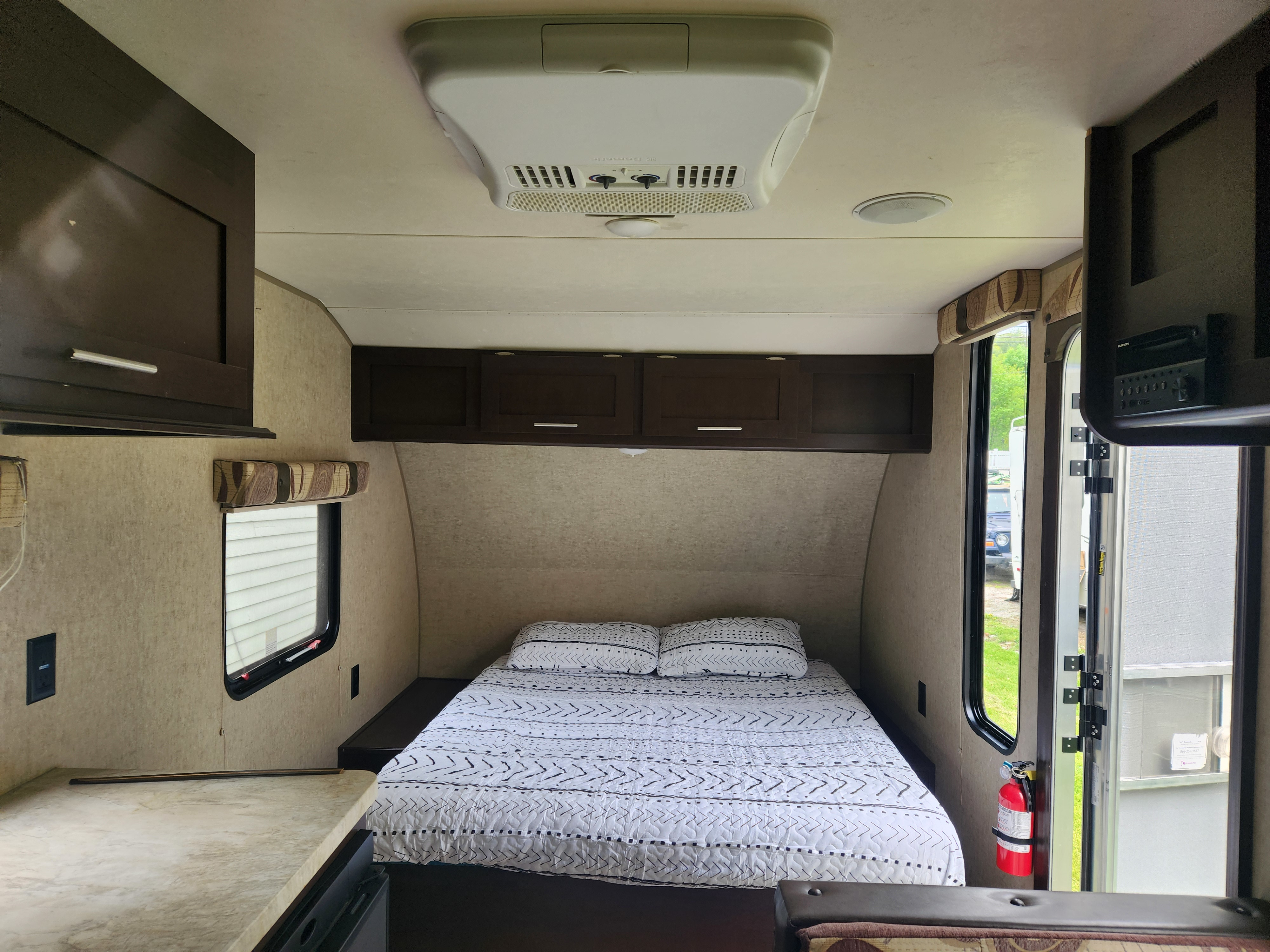Cozy rv rental bedroom: patterned bed, overhead cabinets, AC unit, window view, and fire extinguisher near the door.. Forest River Cherokee Wolf Pup 2017