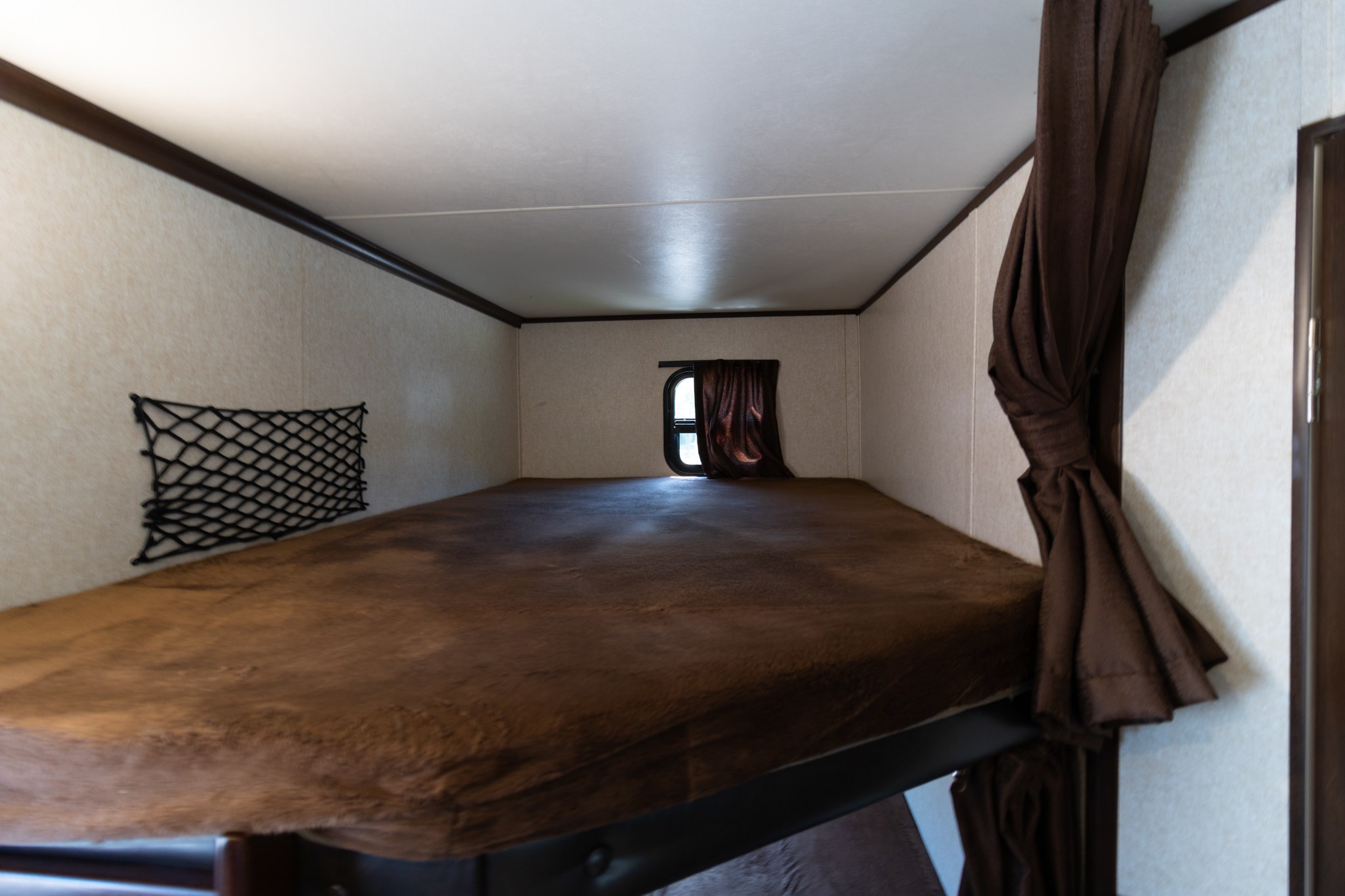 This RV rental bunk bed features a brown mattress, a small window with a curtain, and a storage net for a comfortable sleeping area.. Jayco Jay Flight 2019