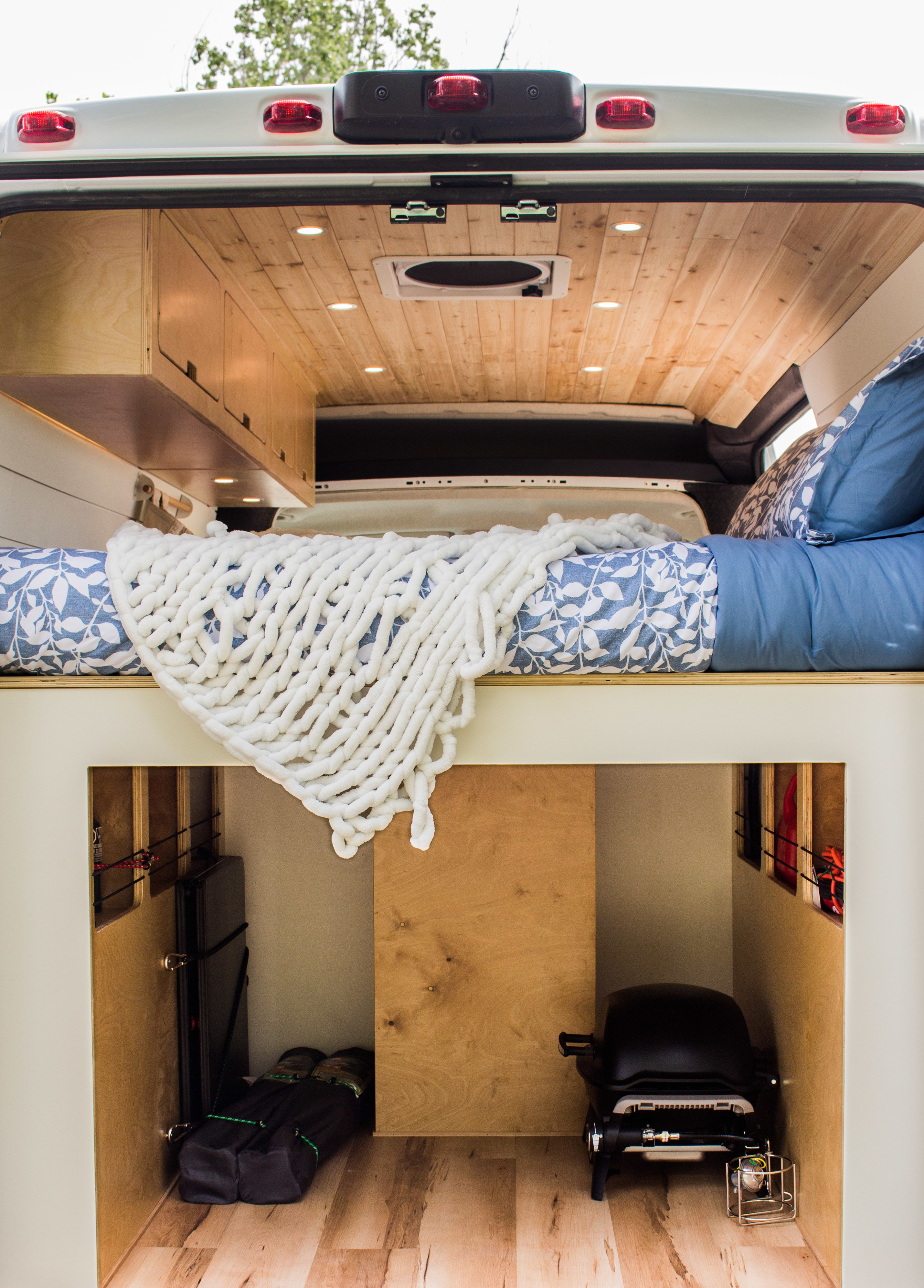 Cozy RV rental interior with bed & chunky blanket, overhead cabinets, and organized rear storage featuring a portable grill.. Ram Promaster 2025