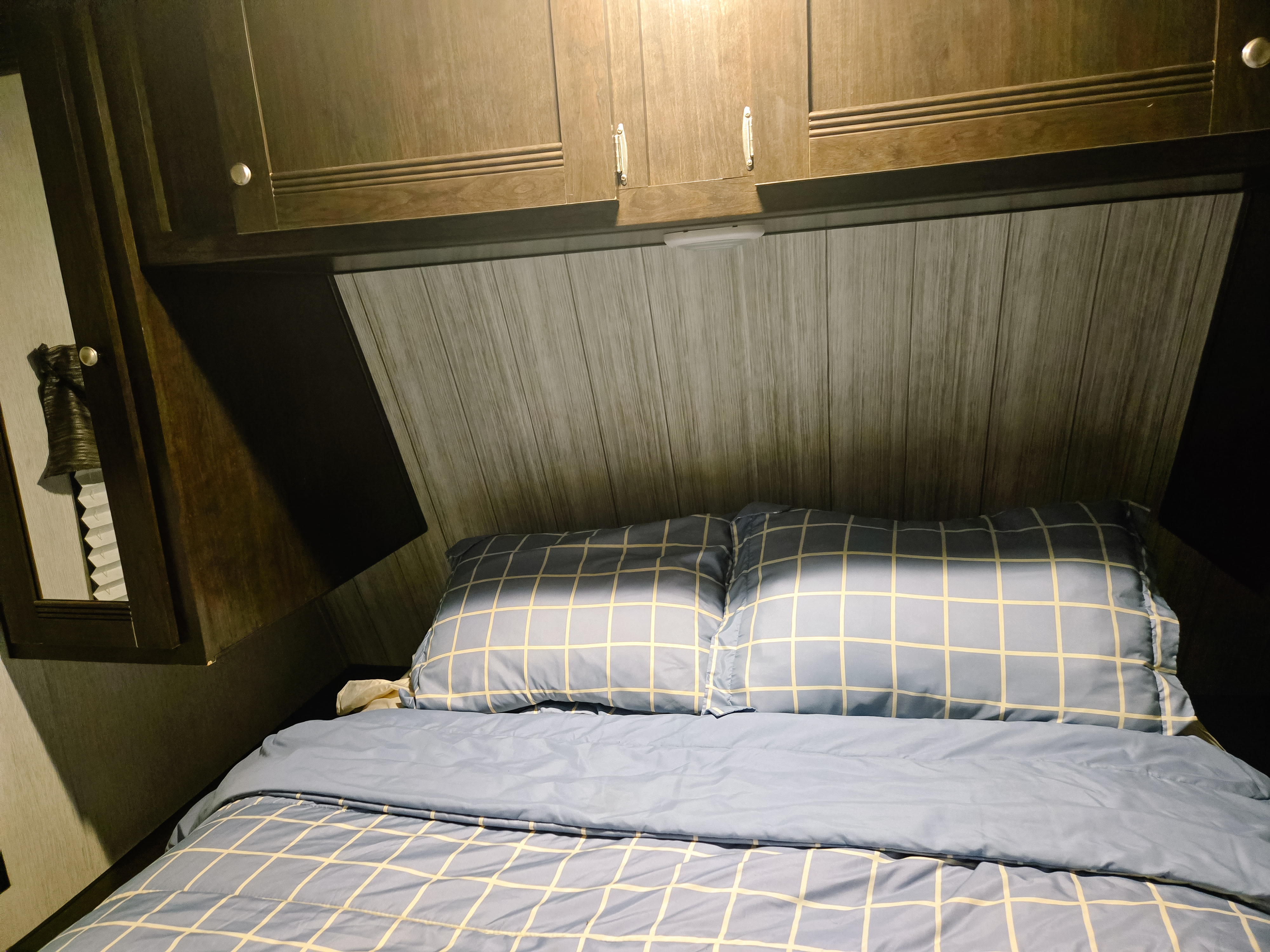 This RV rental shows a compact bedroom with blue checkered bedding under dark wood cabinets and a paneled headboard.. Heartland RVs Pioneer 2019