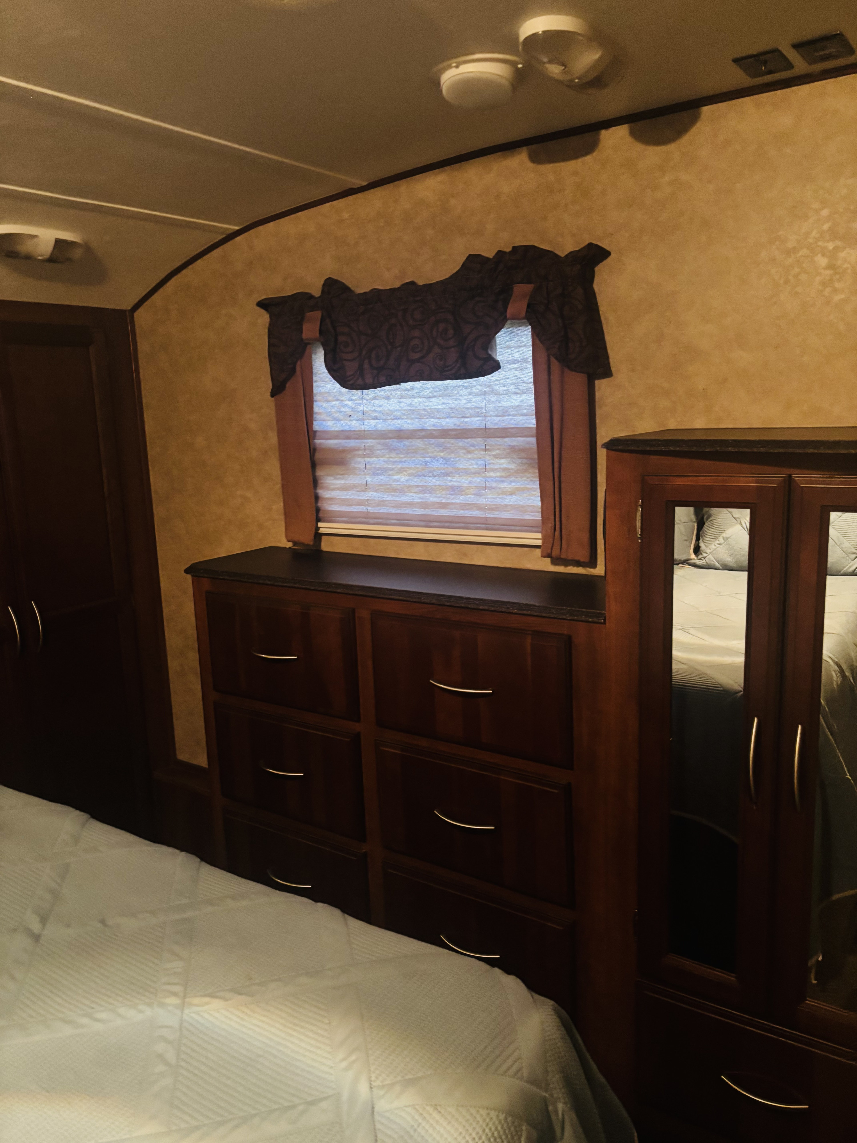 Cozy RV rental bedroom with a bed, wooden dresser, mirrored wardrobe, and a window with blinds and valance.. Forest River 360bhs 2013