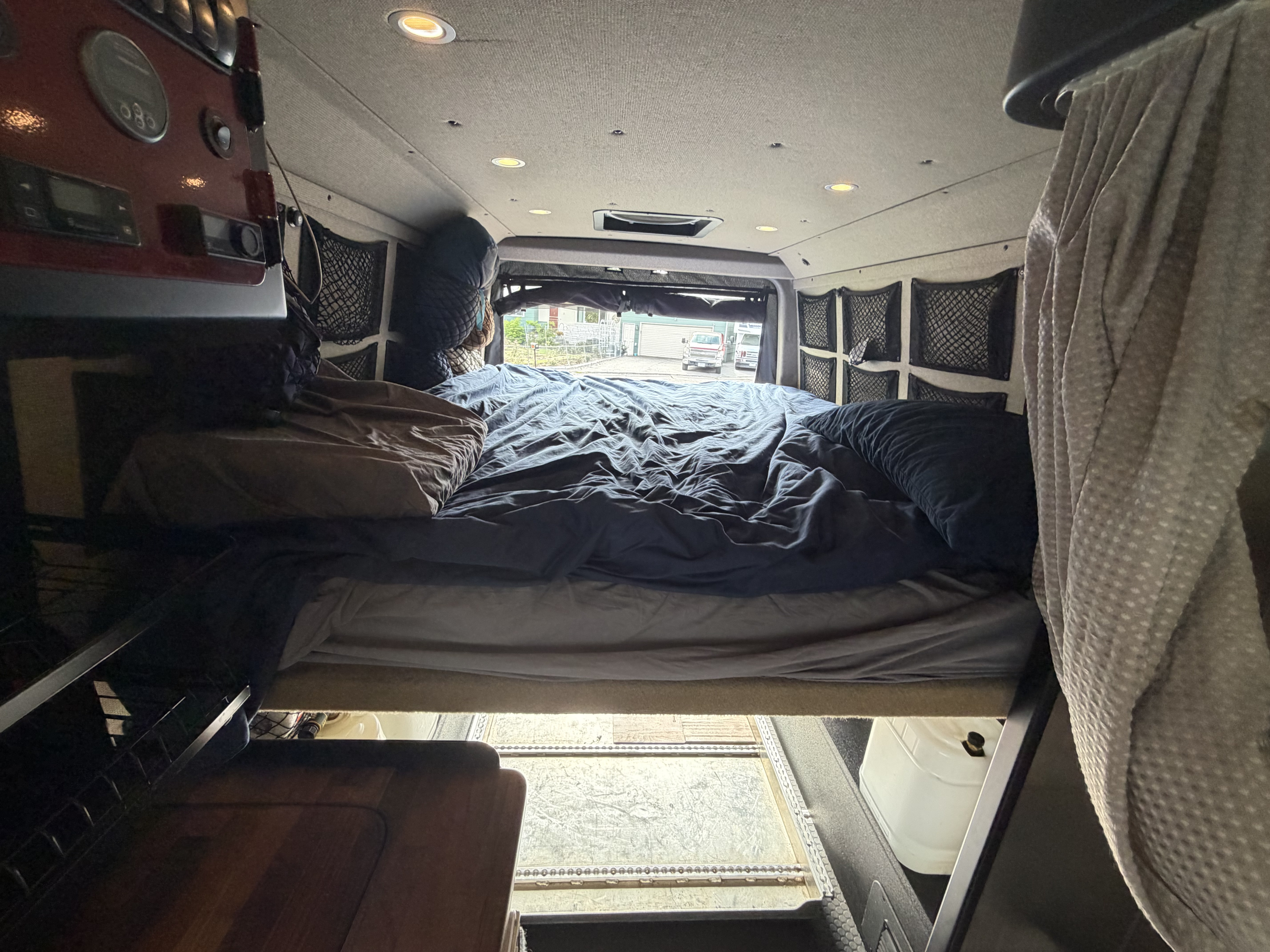 Inside a comfy RV rental, featuring a van bed with blue sheets, overhead lights, and practical side storage nets.. Mercedes Sprinter 2019