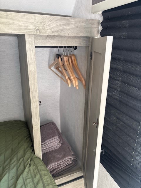 RV rental wardrobe: open closet with wooden hangers, folded towels, and a bed with green bedding. Black blinds are visible.. Sunset Park RV Sun Lite LTD 13MJ 2025