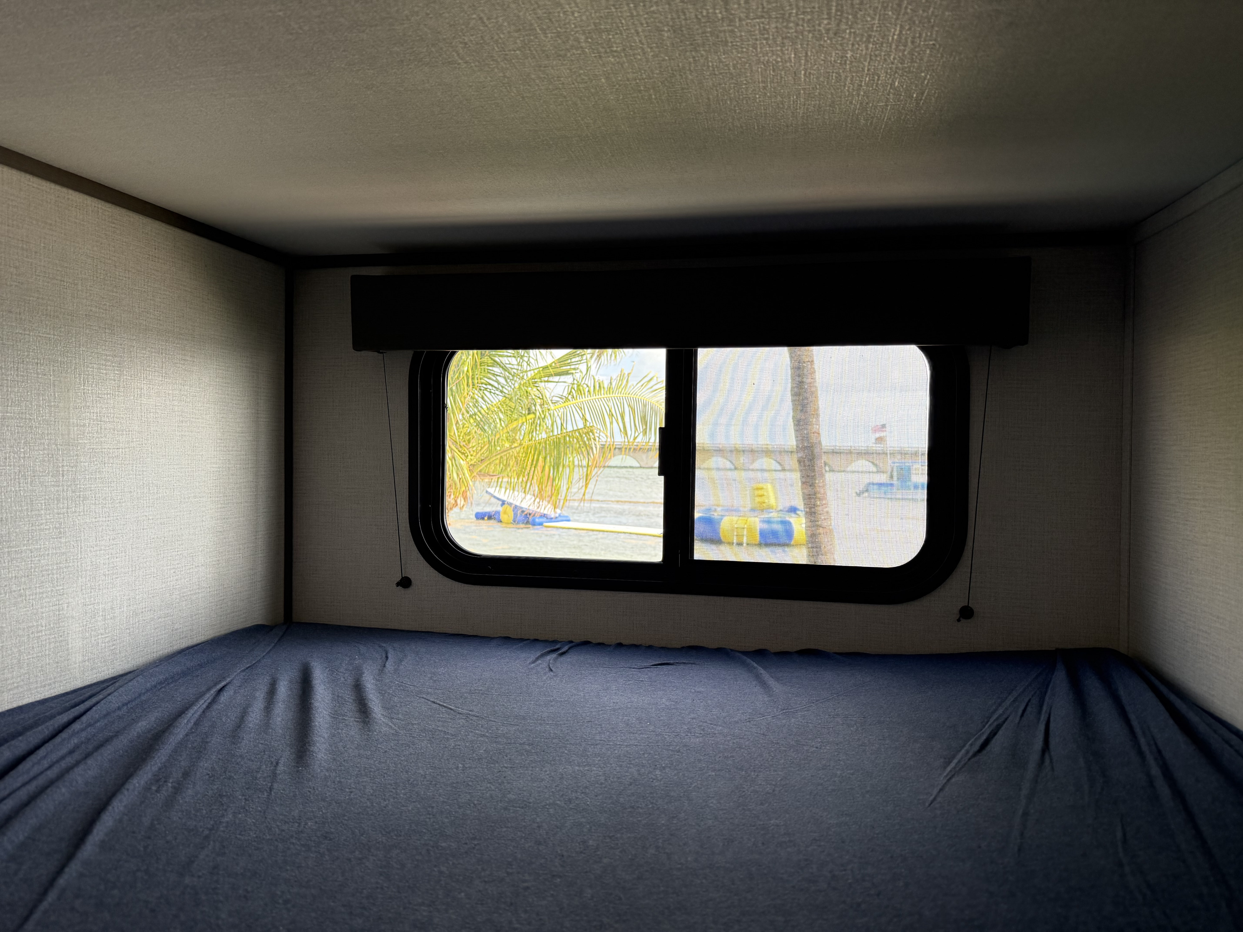 Inside an RV rental: a bunk bed with blue sheets and a window revealing palm trees, water, and a bridge.. Grand Design Transcend Xplor 2026