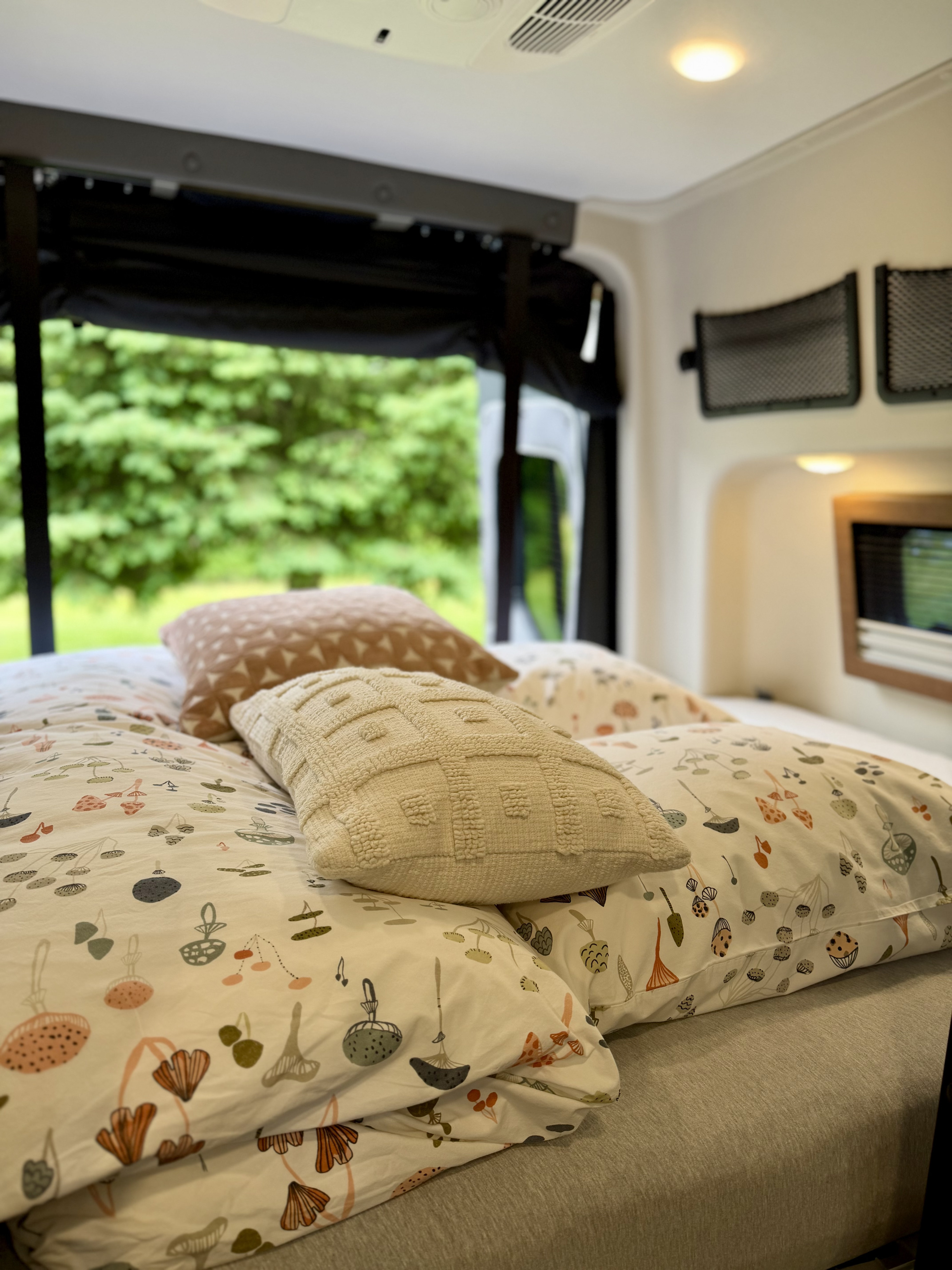 A cozy rv rental bedroom with mushroom-patterned bedding & textured pillows. Lush green trees are visible outside the window.. Winnebago Revel 2025