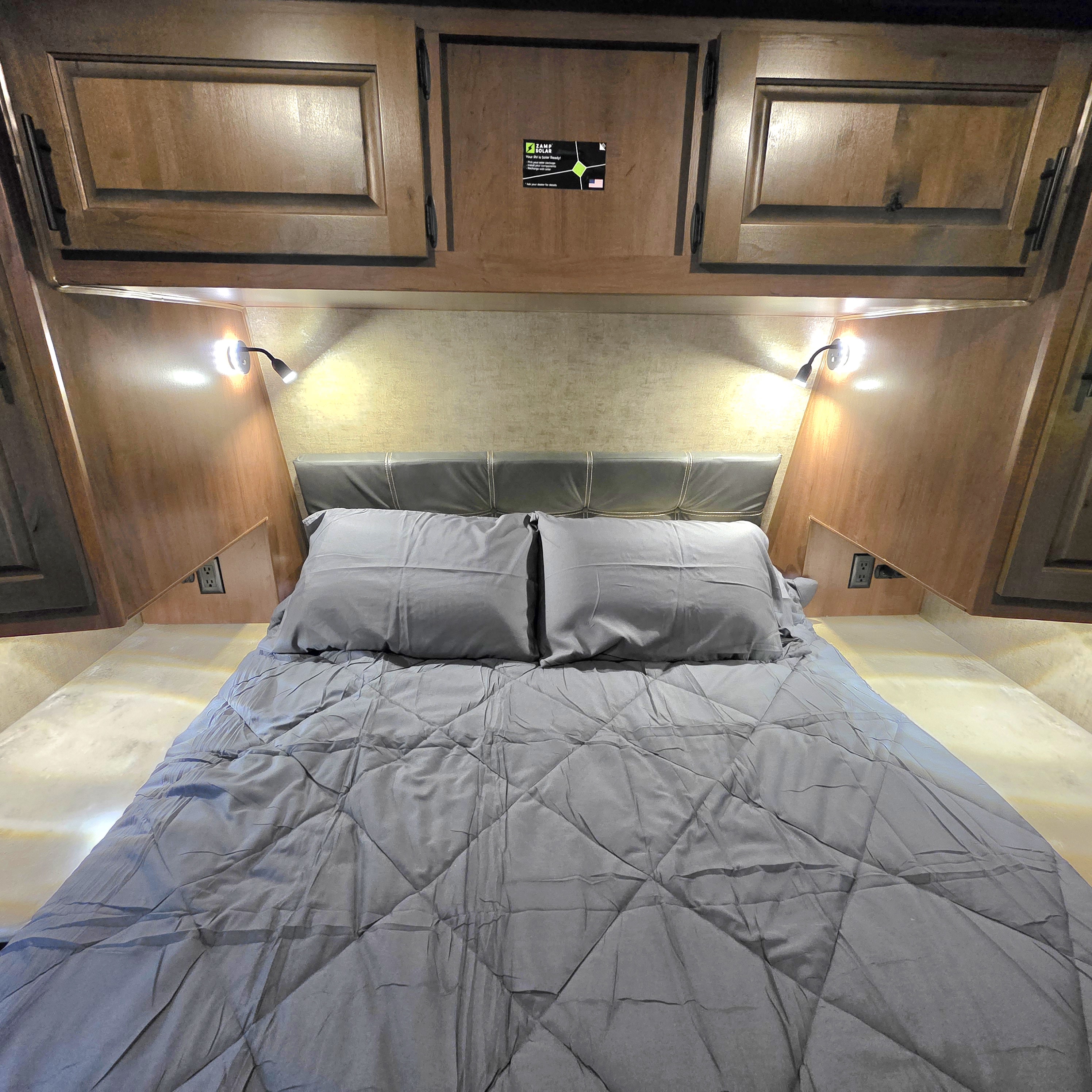 Cozy RV rental bedroom with a bed, gray quilted bedding, a dark headboard, wooden cabinets, and bright reading lights.. Outdoors RV 23DBS 2023