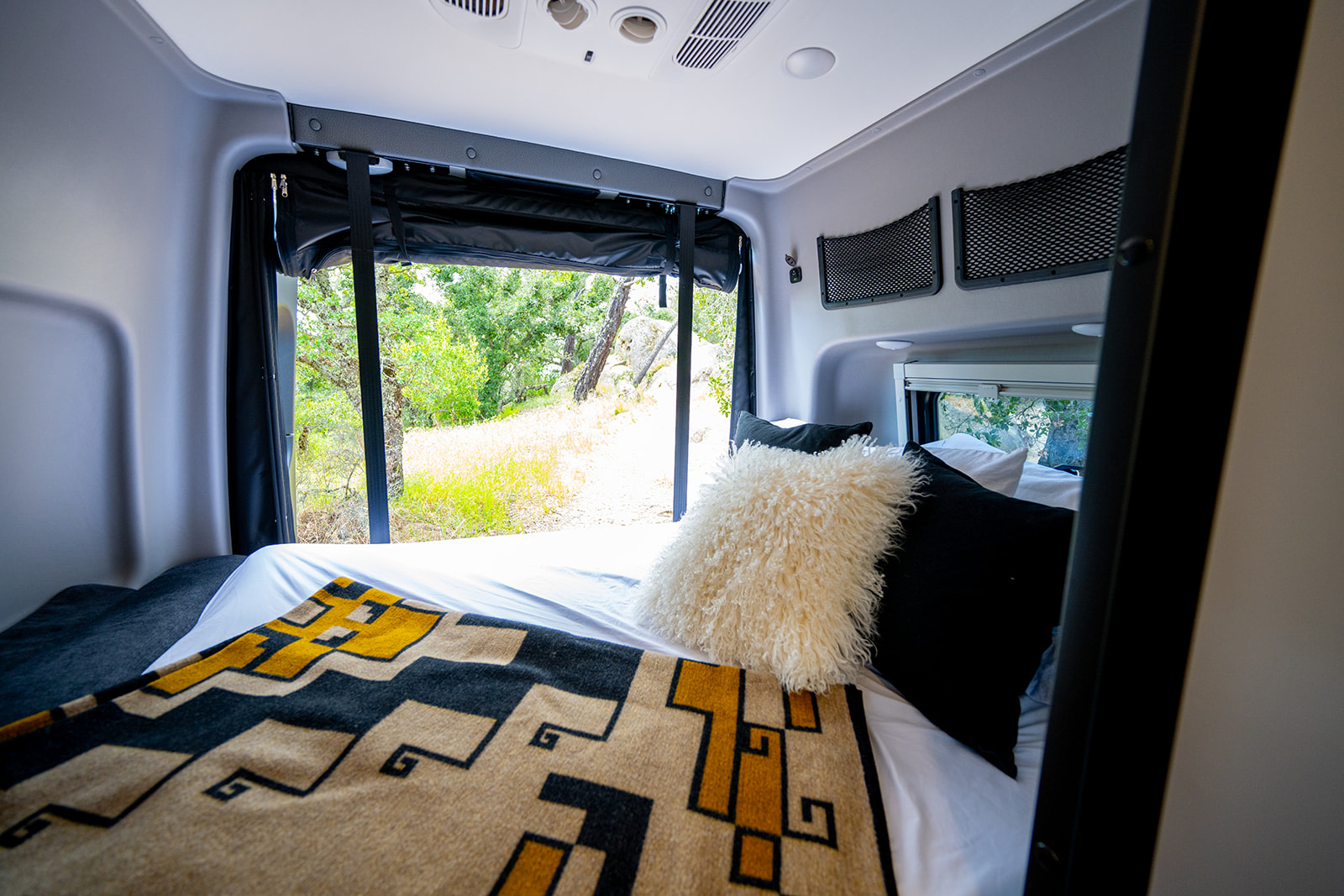 Bedroom of an rv rental showcasing a cozy bed with decorative blanket, scenic window view, and practical overhead storage nets.. Winnebago Revel 2026