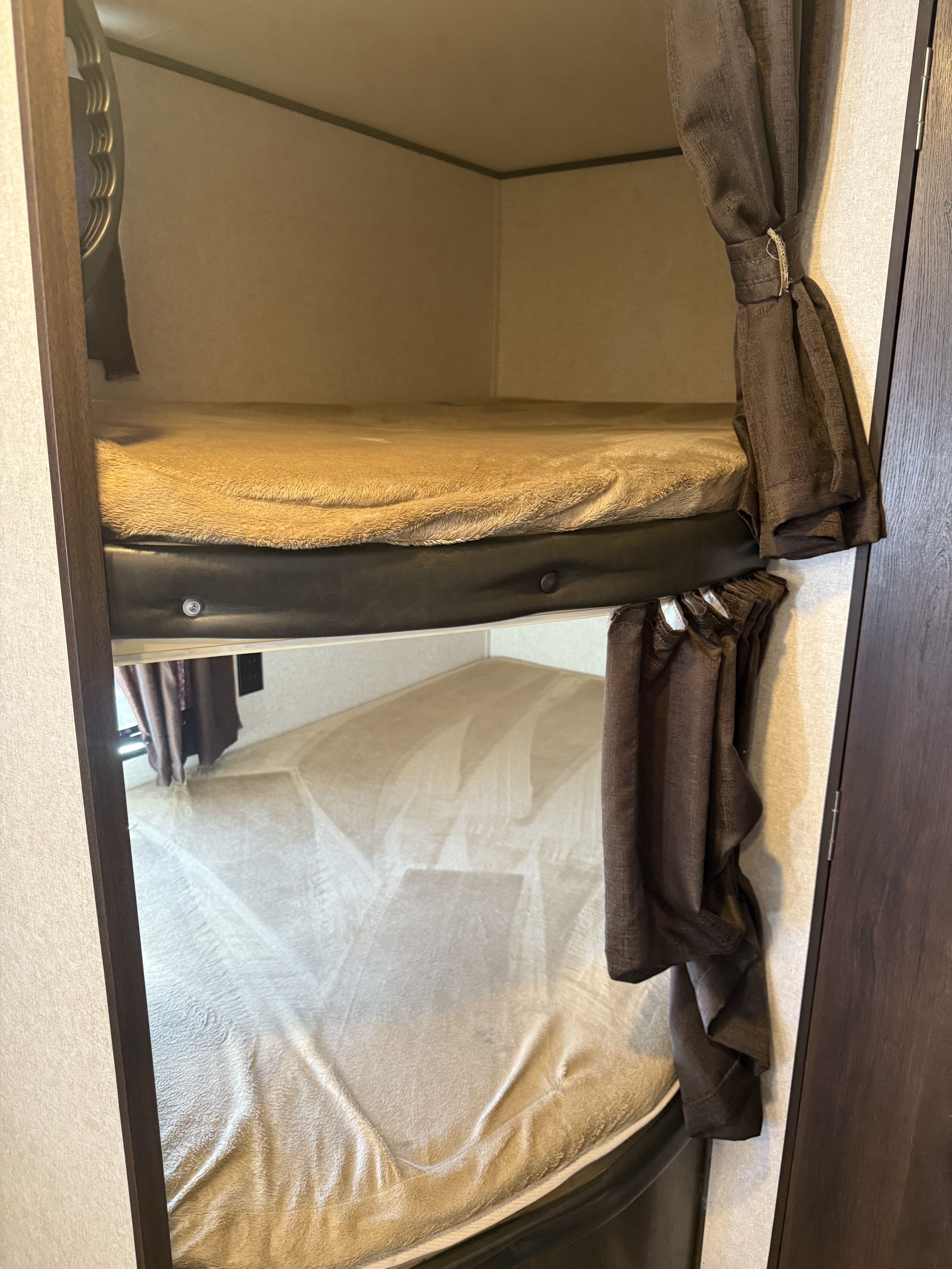 Inside an RV rental, stacked bunk beds are ready with soft blankets. Dark privacy curtains are tied back, next to a wood door.. Jayco Jay Flight SLX Baja Edition 2018