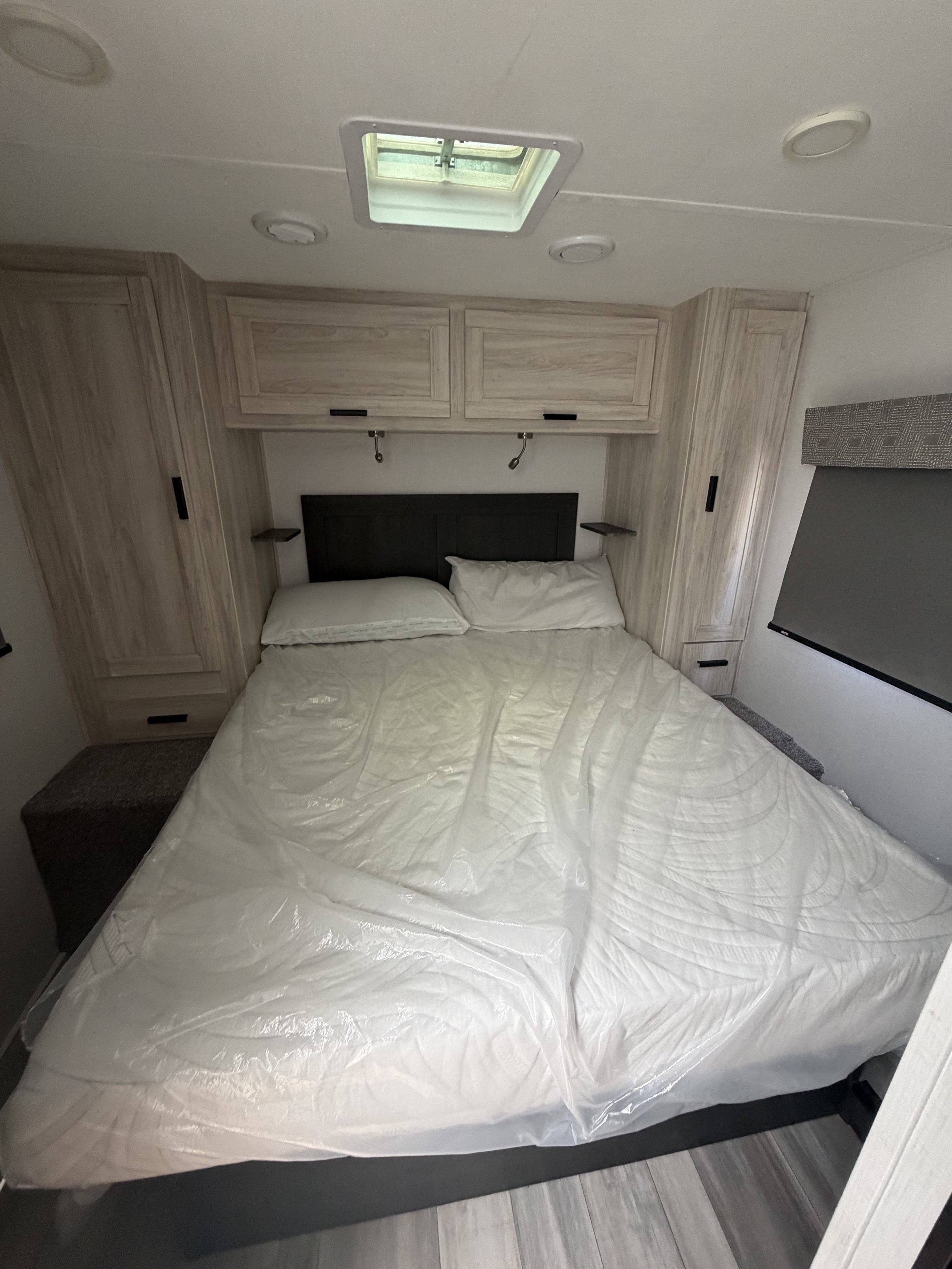 RV rental bedroom: New bed (in plastic), dark headboard, wood cabinets, skylight, and reading lights for comfortable travel.. Forest River Sunseeker LE 2024