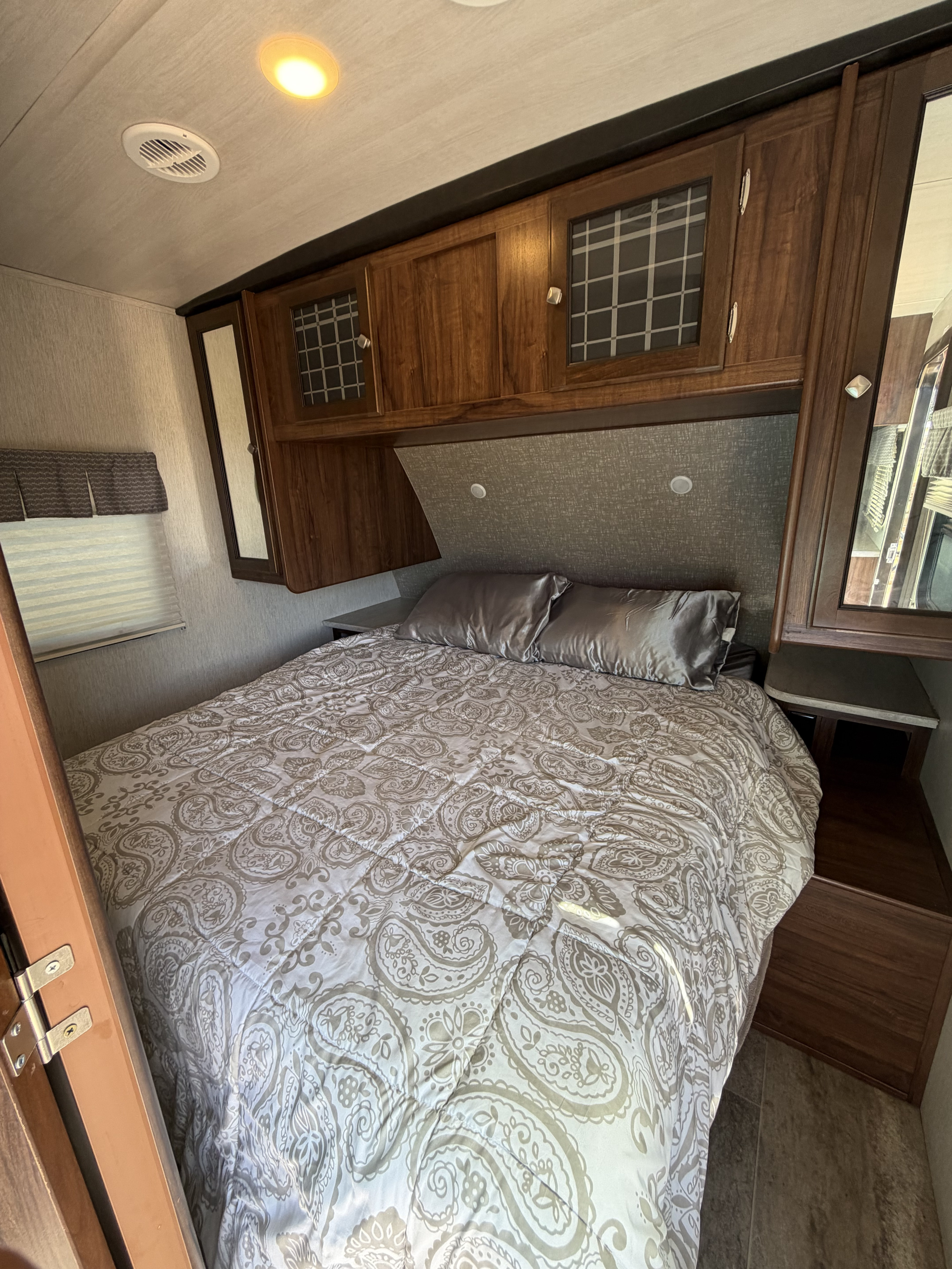 RV rental bedroom: cozy bed with a patterned comforter, satin pillows, wood overhead cabinets, and a window with blinds.. Heartland RVs Wilderness 2018