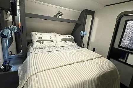 Cozy RV rental bedroom with striped bedding, camper-themed pillows, and a window, ready for your next travel adventure.. Dutchmen Coleman Lantern 2022
