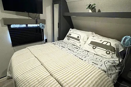 Cozy RV rental bedroom: bed with striped comforter & adventure-themed pillows. TV, window, shelf, and small fans visible.. Dutchmen Coleman Lantern 2022