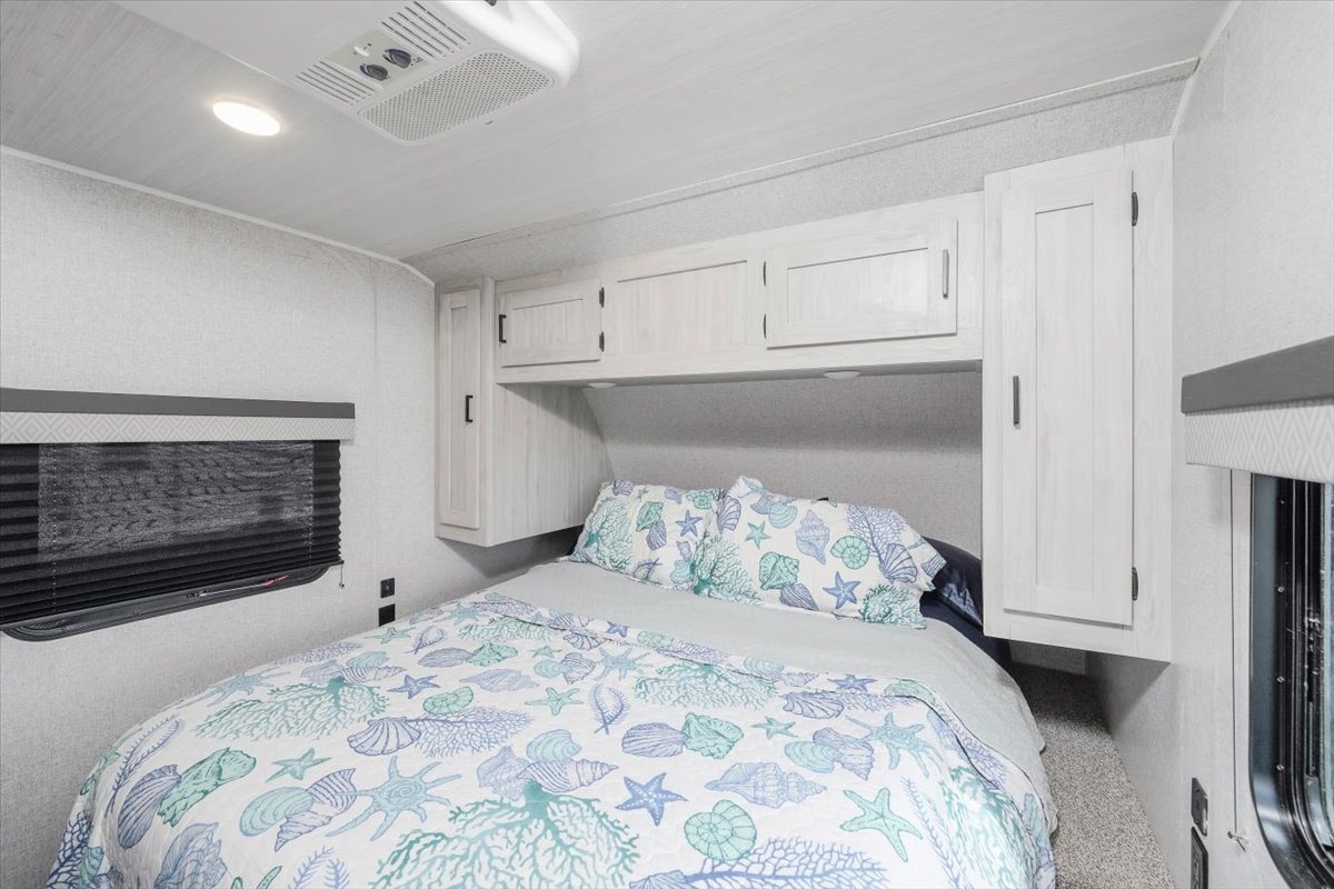 This cozy RV rental bedroom features a queen bed with seashell and coral bedding, overhead storage cabinets, and a window with blinds.. East to West Della Terra 2023