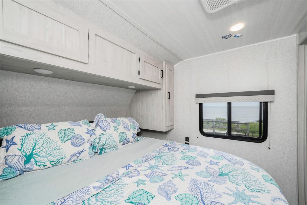 This RV rental bedroom features a comfy bed with marine-themed bedding, overhead cabinets, and a window offering an ocean view.. East to West Della Terra 2023