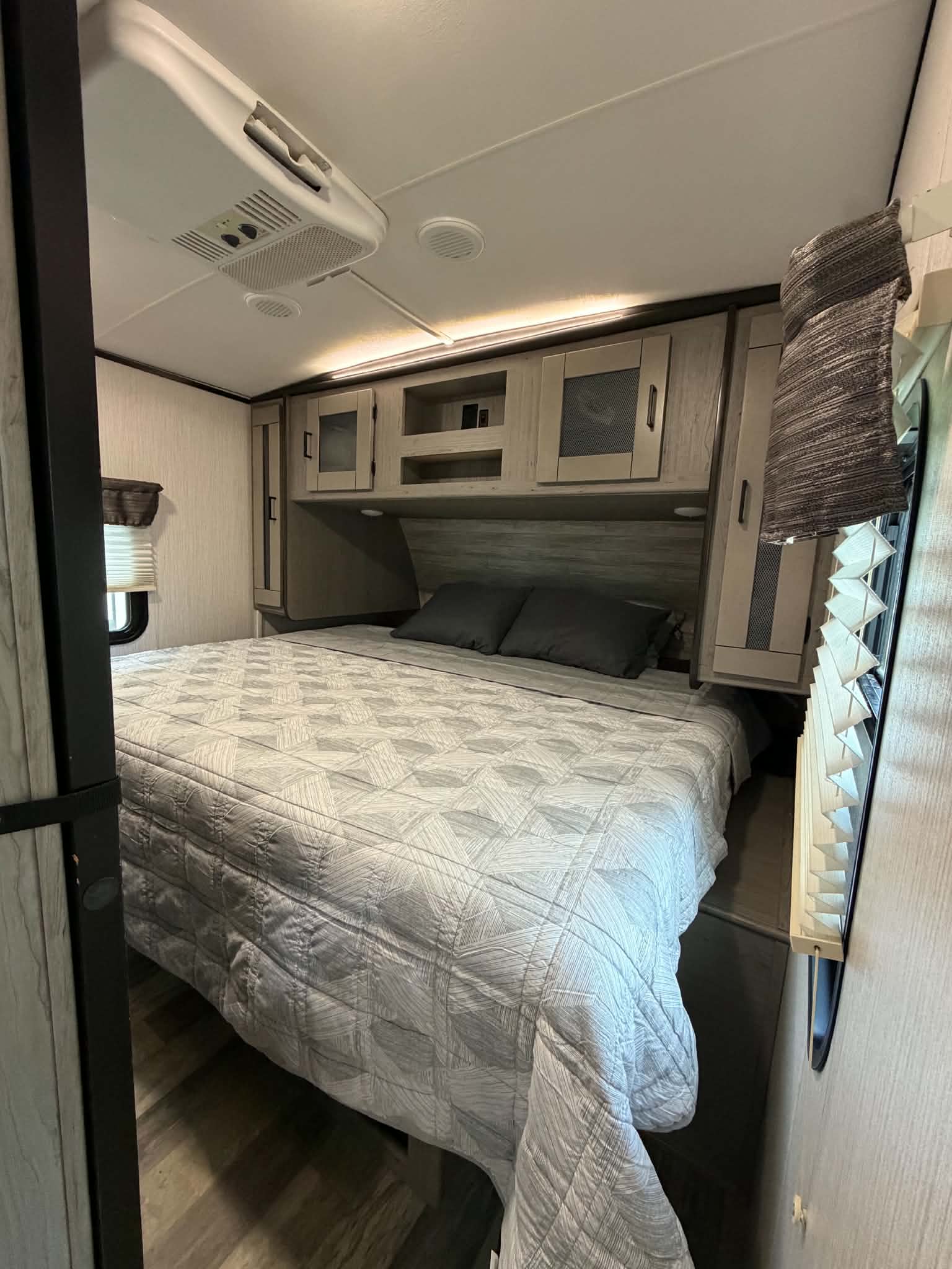 RV rental bedroom with a light grey patterned bedspread, dark pillows, wood-look cabinets, and an AC unit for comfort.. Heartland RVs Mallard 2020