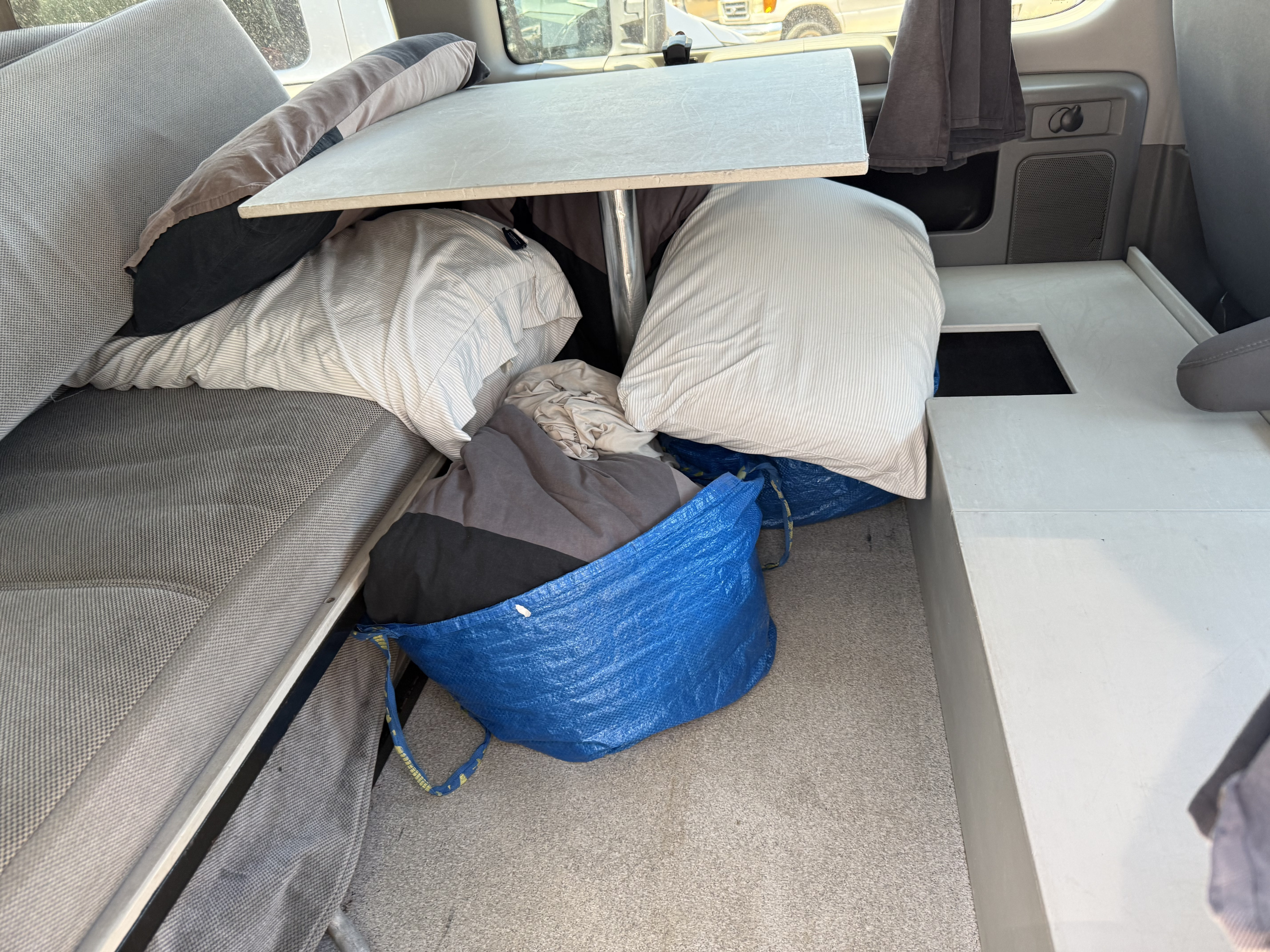 Inside this rv rental, a compact living area features a bed, dining table, and personal belongings stored in blue bags.. Ford E350 Box Van 2013