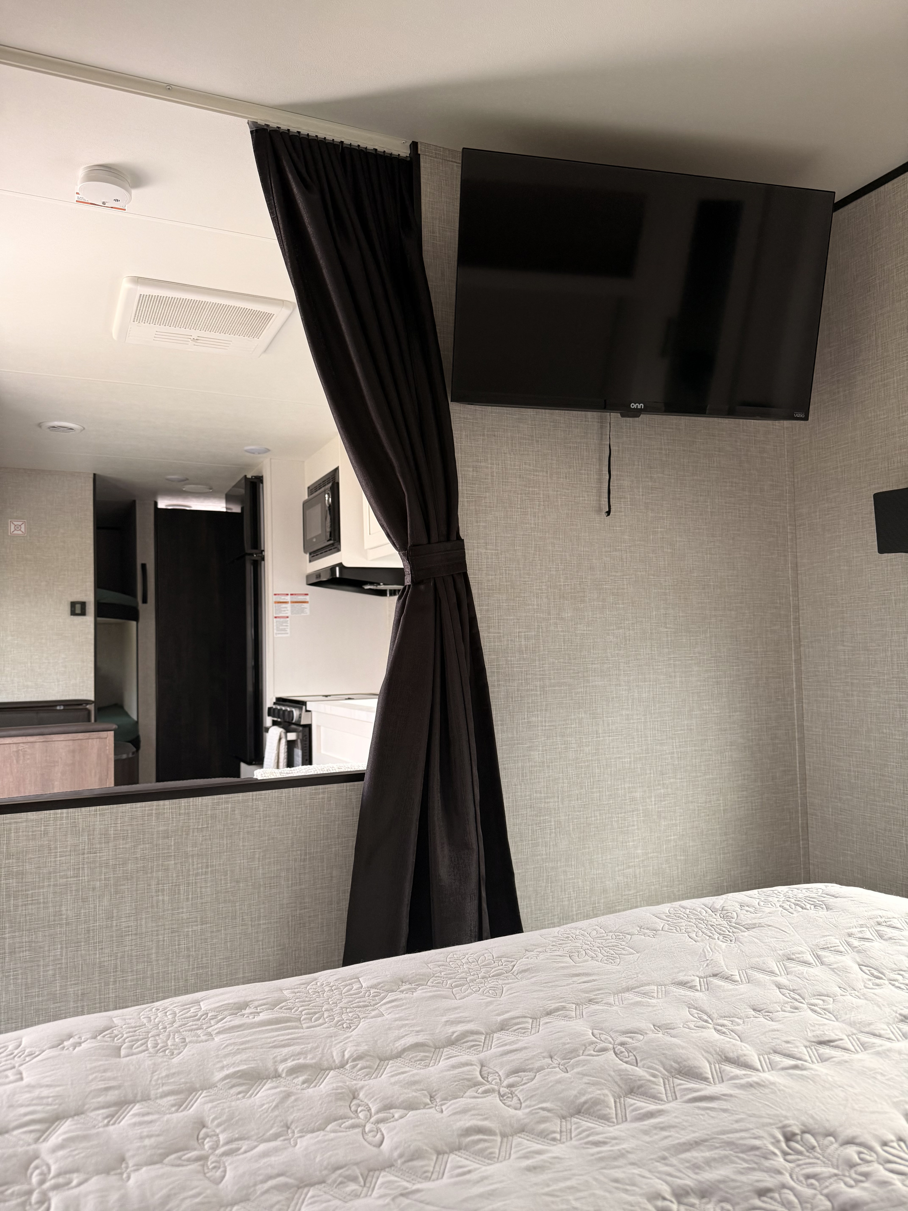 rv rental interior: Bedroom with white quilted bed, TV, dark privacy curtain, and open view to compact kitchen.. Jayco Jay Flight SLX 2024