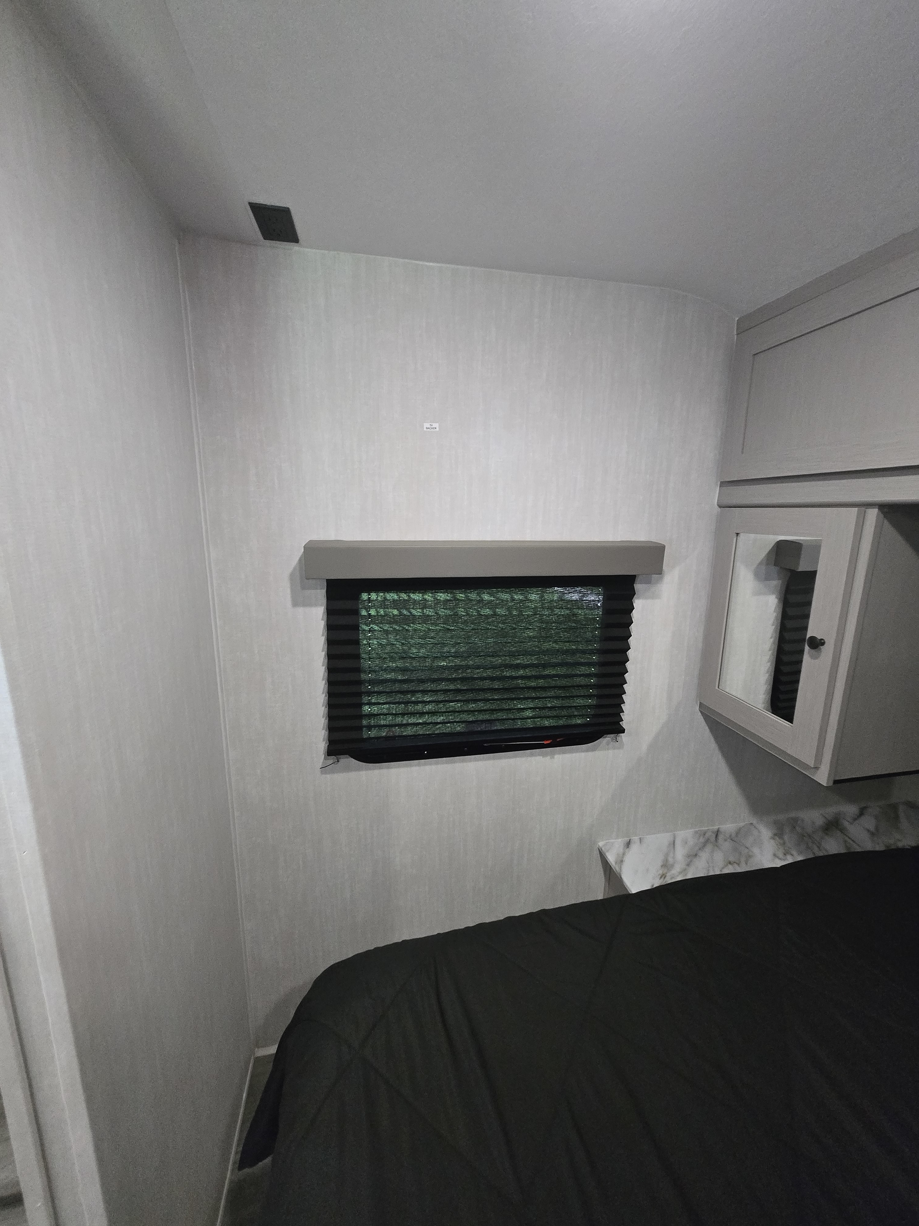 Cozy RV rental bedroom featuring a window with dark blinds, a bed with a black comforter, overhead cabinets, and a TV mount area.. Coachmen Apex X 2025