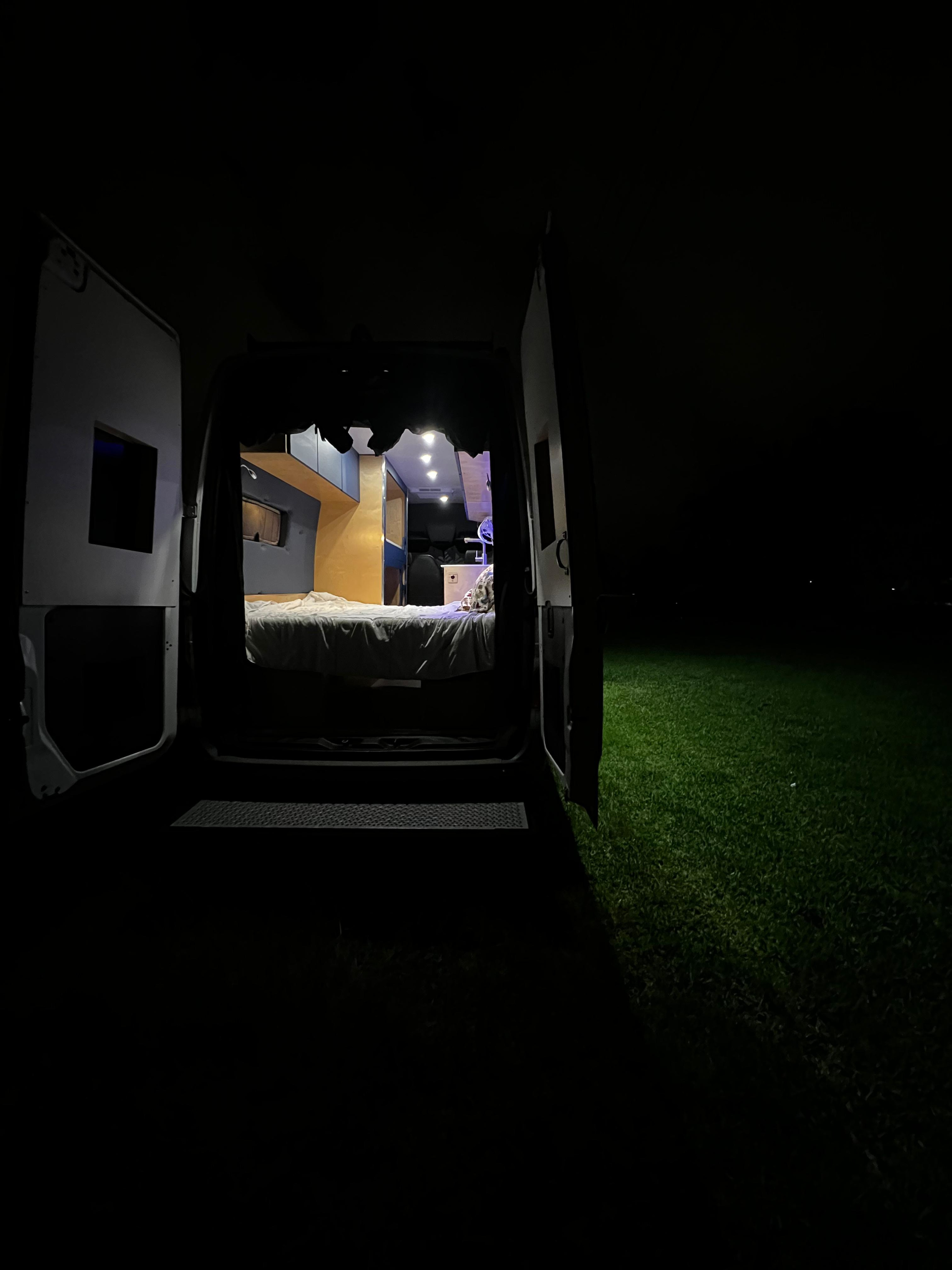 Cozy rv rental camper van interior at night with open doors, illuminating grass outside.. Mercedes-Benz Sprinter 2023
