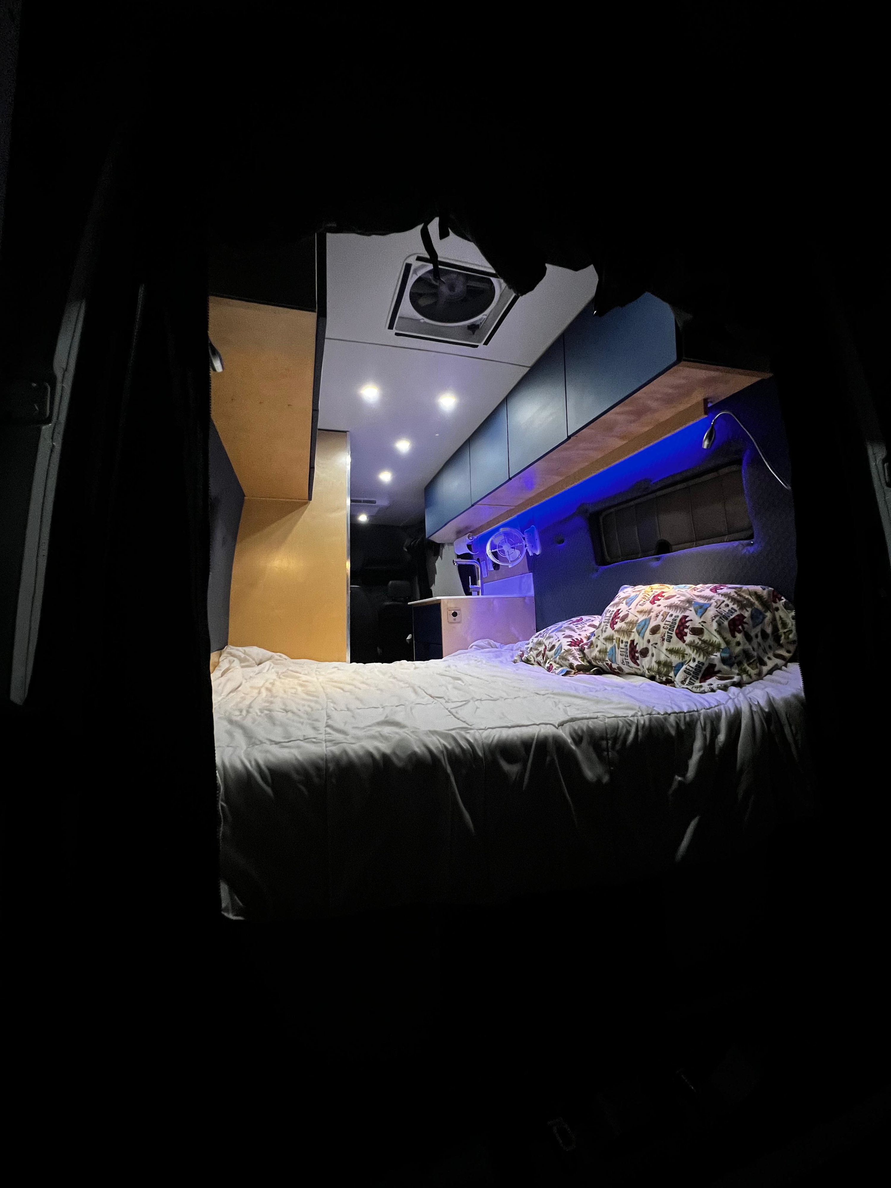 Inside this rv rental, a cozy bed with patterned pillows, blue accent lighting, overhead spotlights, and a ceiling fan create comfort.. Mercedes-Benz Sprinter 2023