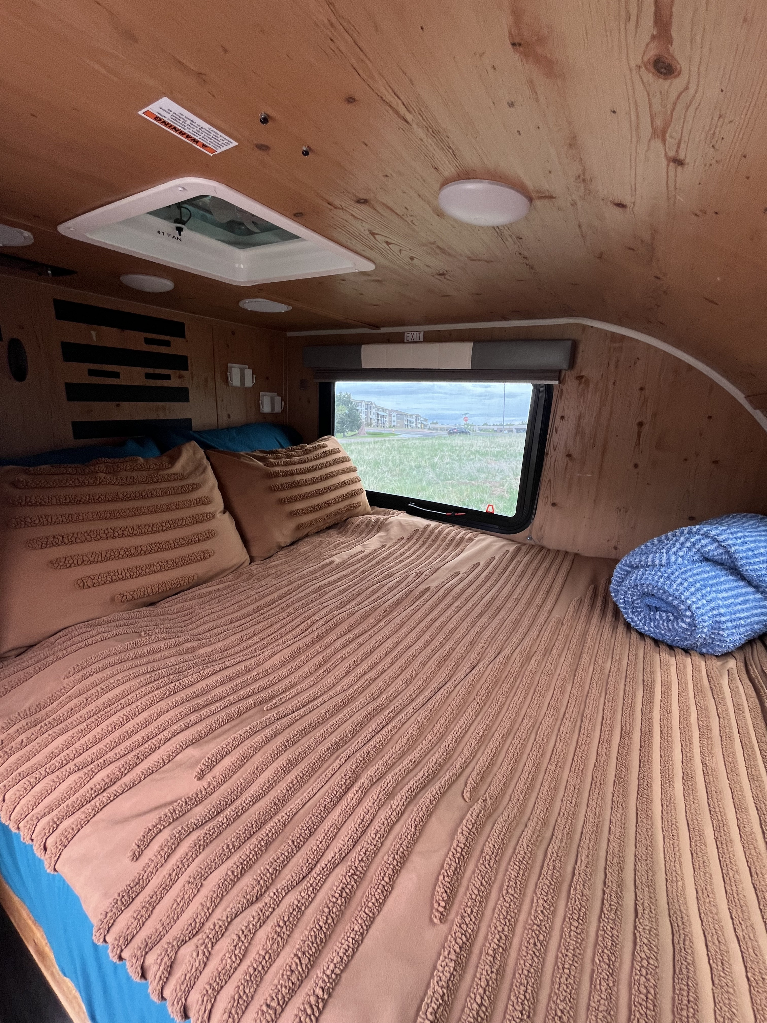 Cozy RV rental sleeping area with wood-paneled walls. Features a textured bed, pillows, and a window view of green fields.. Sunset Park RV SunRay 2021