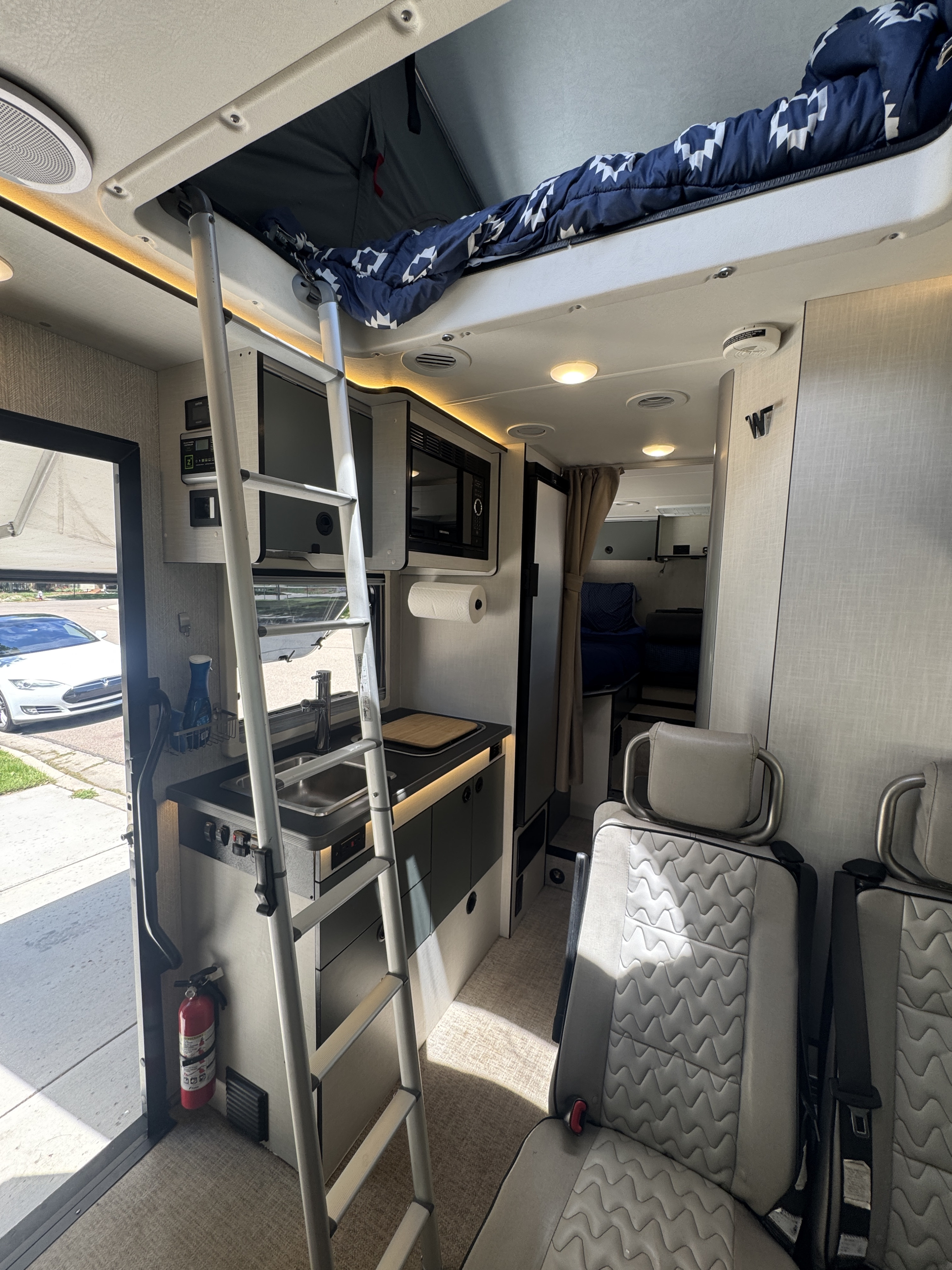RV rental interior showcasing a compact kitchen (sink, microwave), ladder to a bunk bed, and passenger seats. All essentials onboard.. Winnebago Ekko 2022