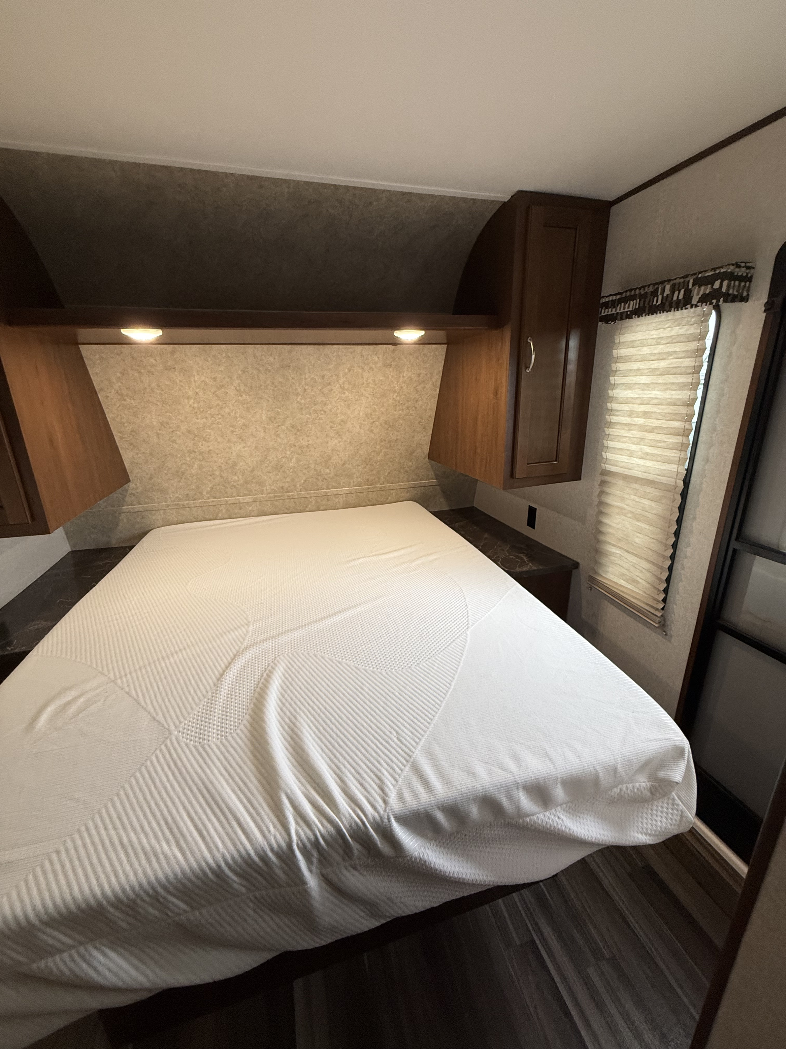 Cozy RV rental bedroom featuring a bed with a white mattress, overhead cabinets, under-cabinet lighting, and a window with blinds.. Jayco Jay Flight SLX 2018