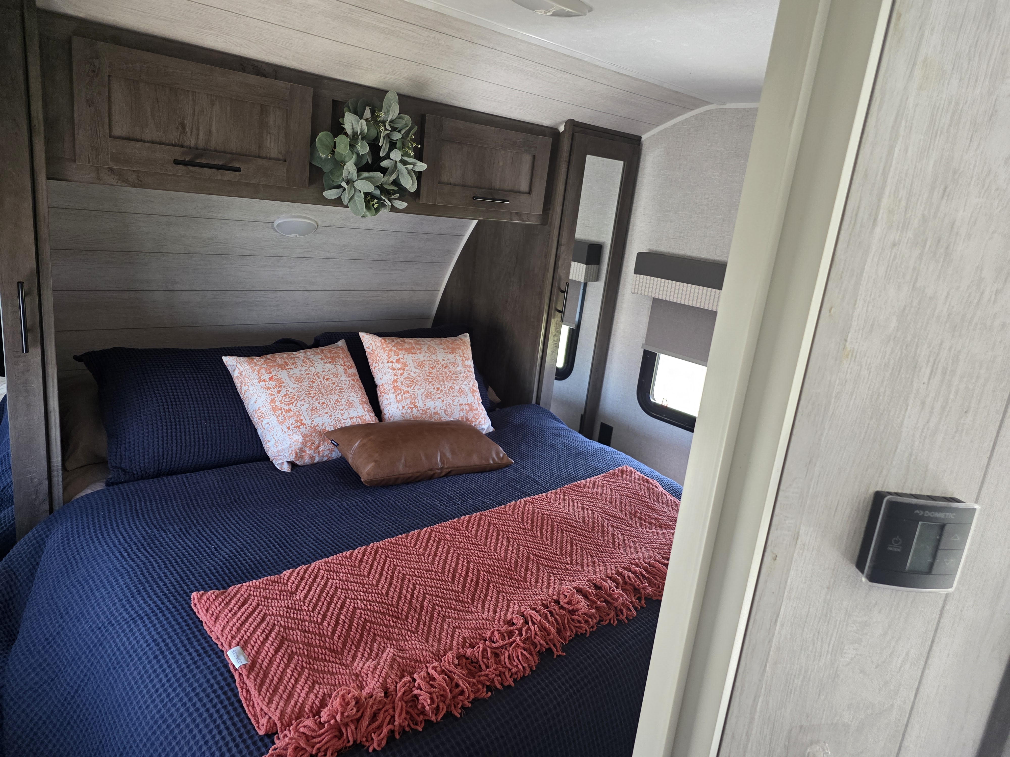Cozy RV rental bedroom with navy bedding, decorative pillows, and a coral throw, featuring rustic cabinetry and windows with shades.. Forest River Wildwood 2021