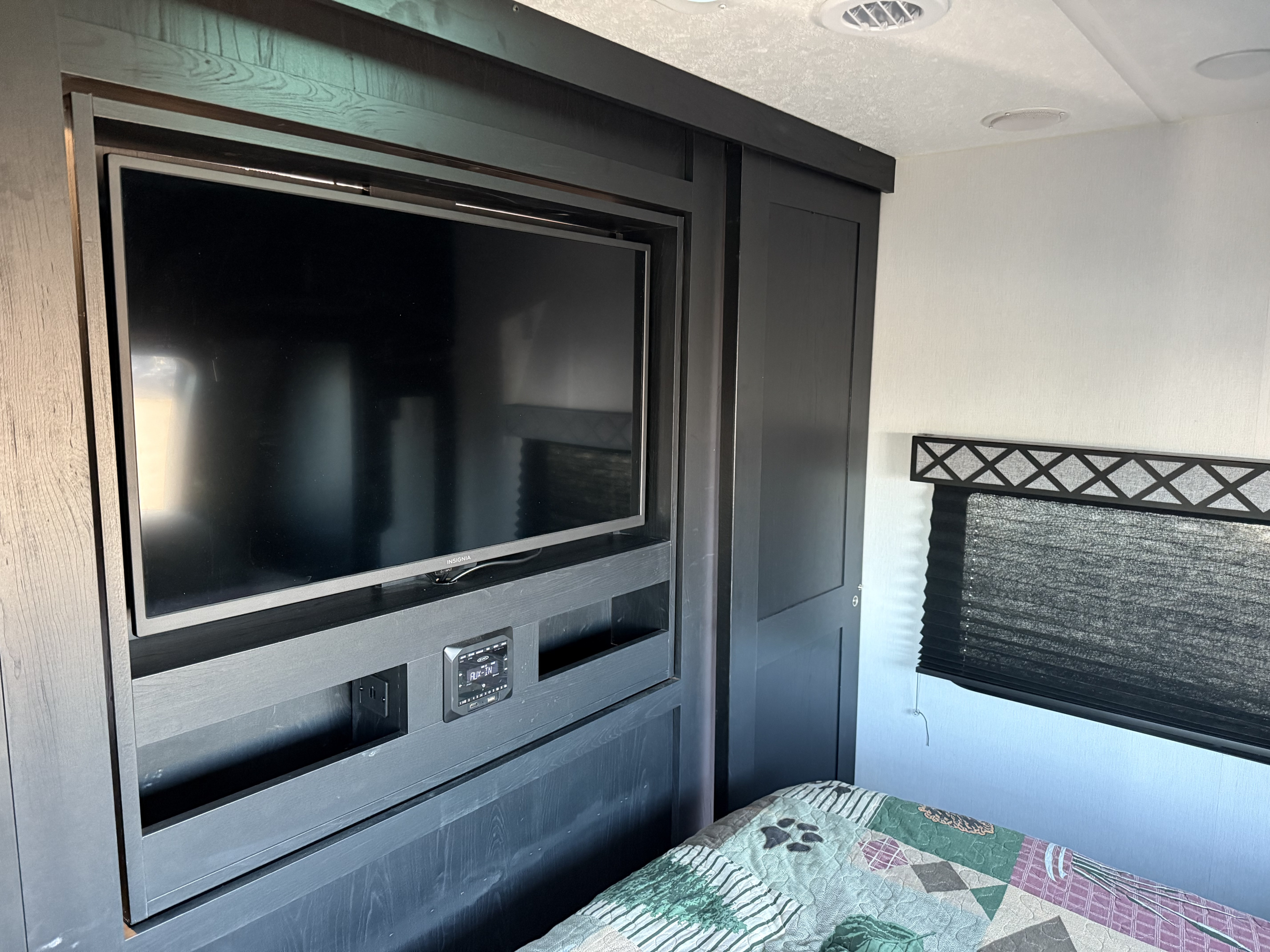 RV rental bedroom interior: dark wood TV/media built-in unit, closet, bed with quilt, and window with pleated blind.. Coachmen Freedom Express 257BHS 2022
