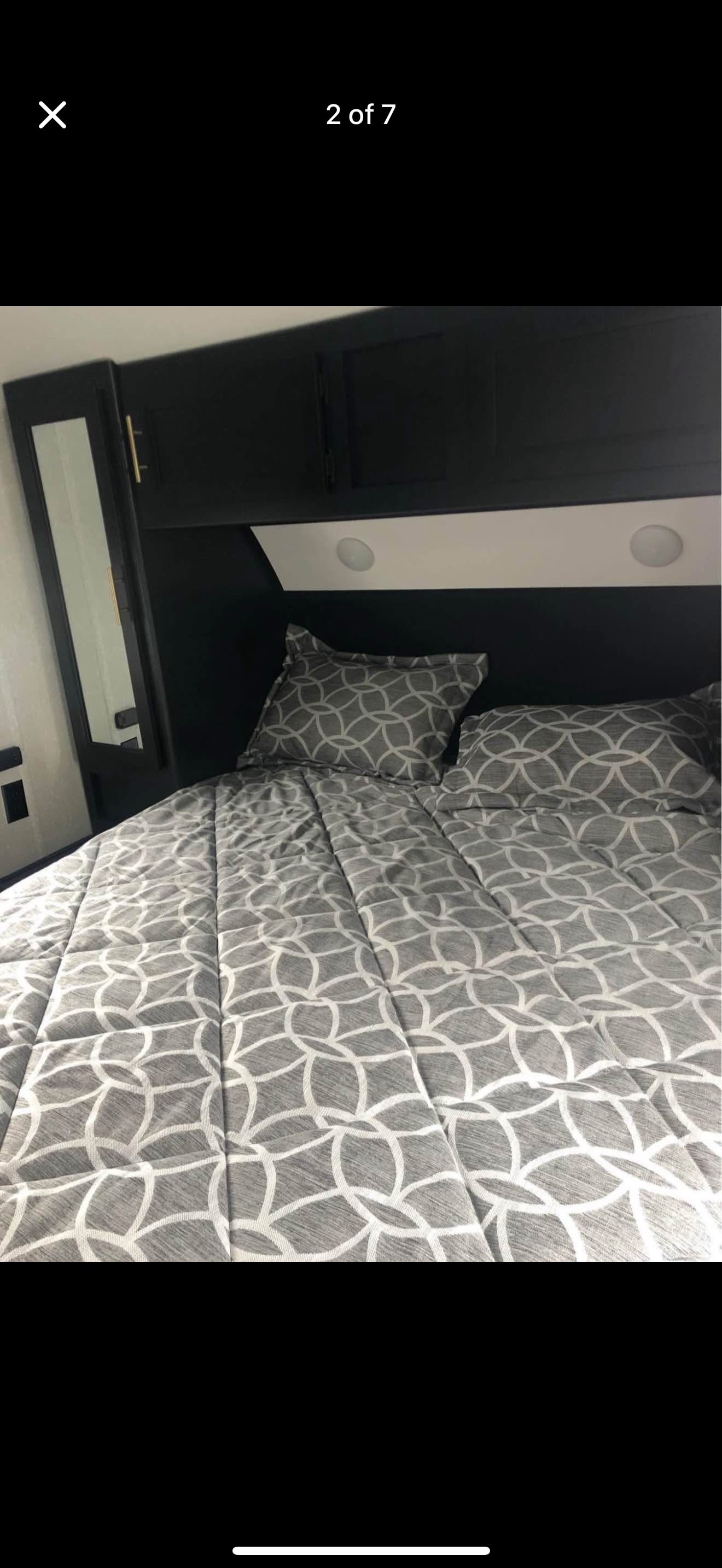 Comfortable bed with grey geometric bedding in an RV rental, complete with dark overhead storage, mirror, and task lighting.. Forest River Cherokee 2022