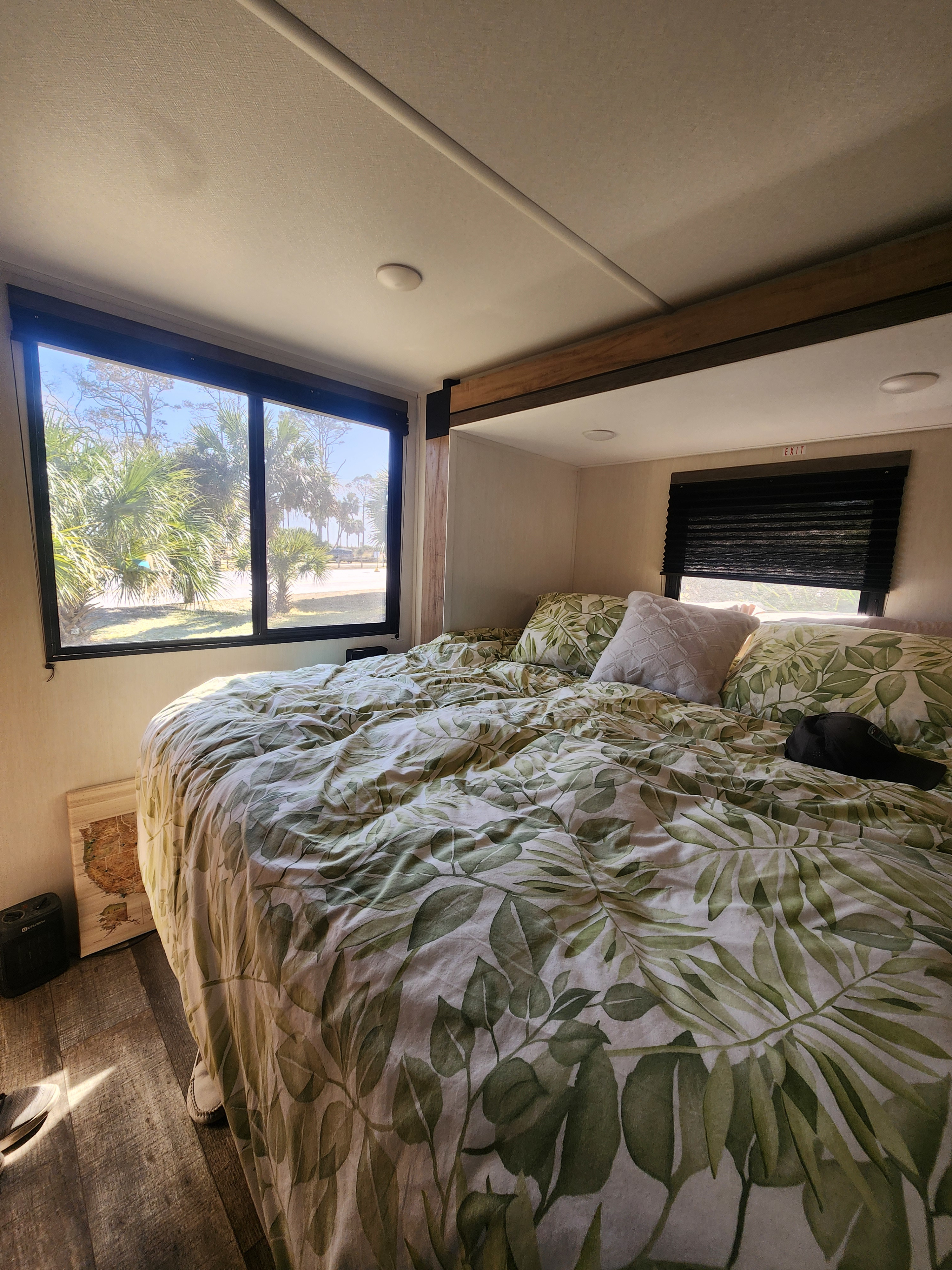 Bright RV rental bedroom features a comfy bed with leaf-patterned duvet, palm tree window view & exit sign.. Forest River Campsite Reserve 2025