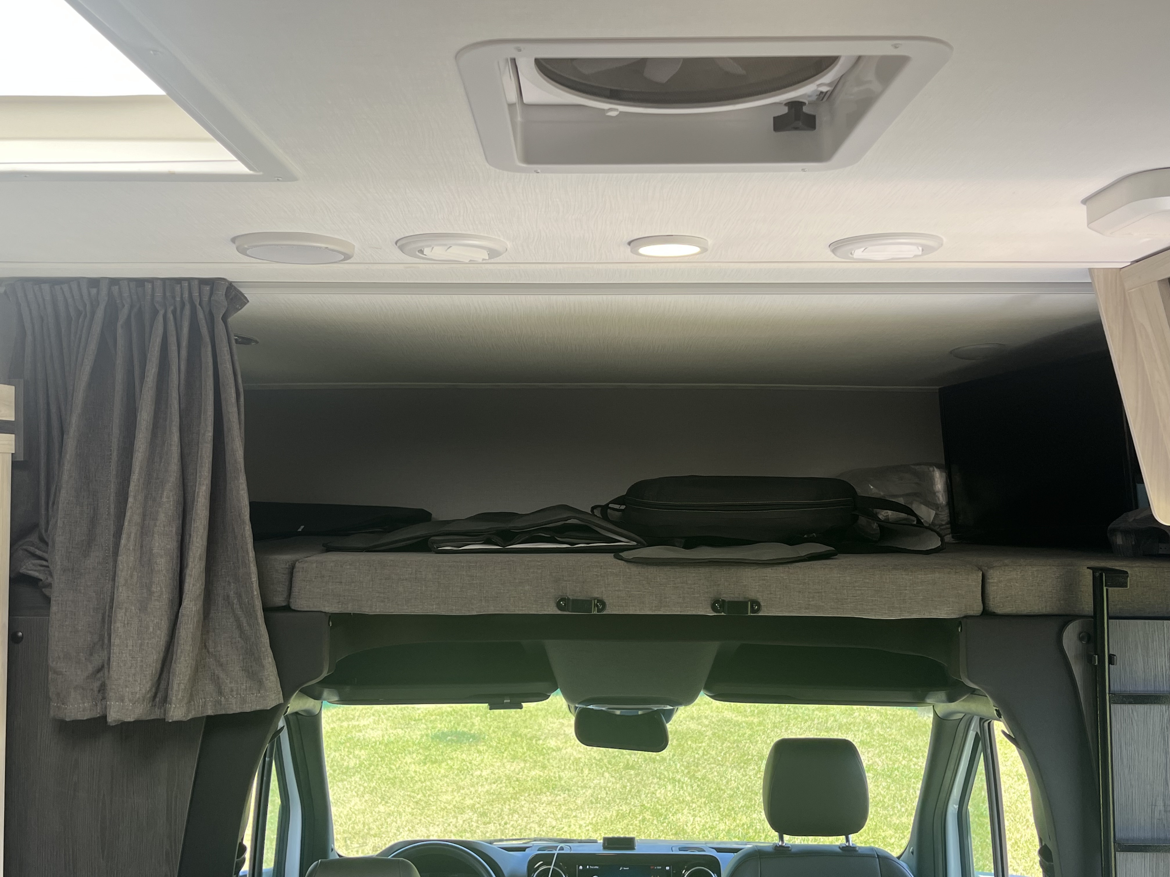 Inside this RV rental: Overhead bed, ceiling with skylight and vent fan, and cockpit view through the windshield to green grass.. Forest River Sunseeker MBS 2023