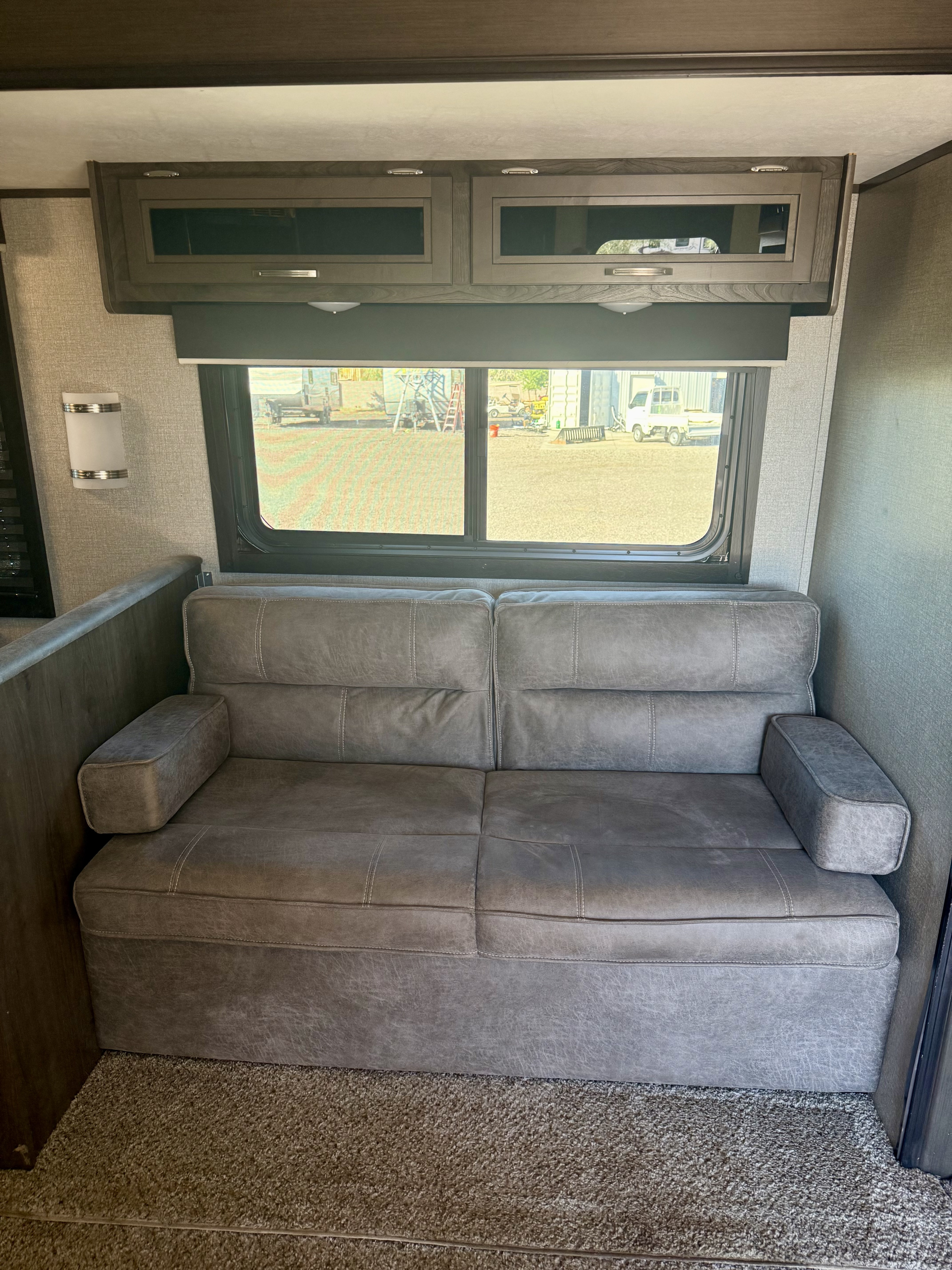 RV rental interior with a comfy gray sofa, overhead cabinets, and a large window with a roll-down shade.. Jayco 267BHSW 2021
