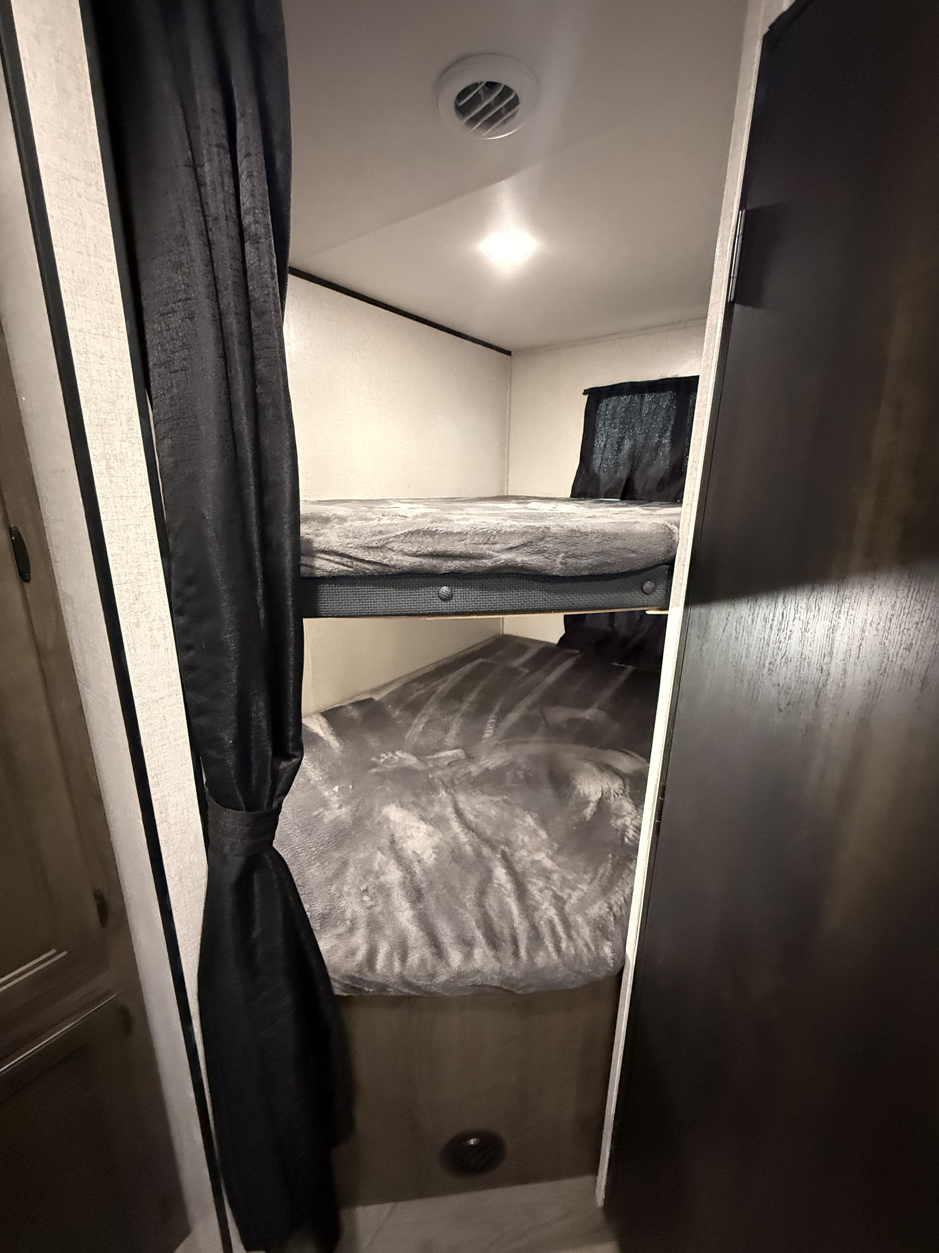 Inside an RV rental: two compact bunk beds with grey bedding, framed by black privacy curtains. Ceiling vent & light above.. Olympia 26BHS 2022