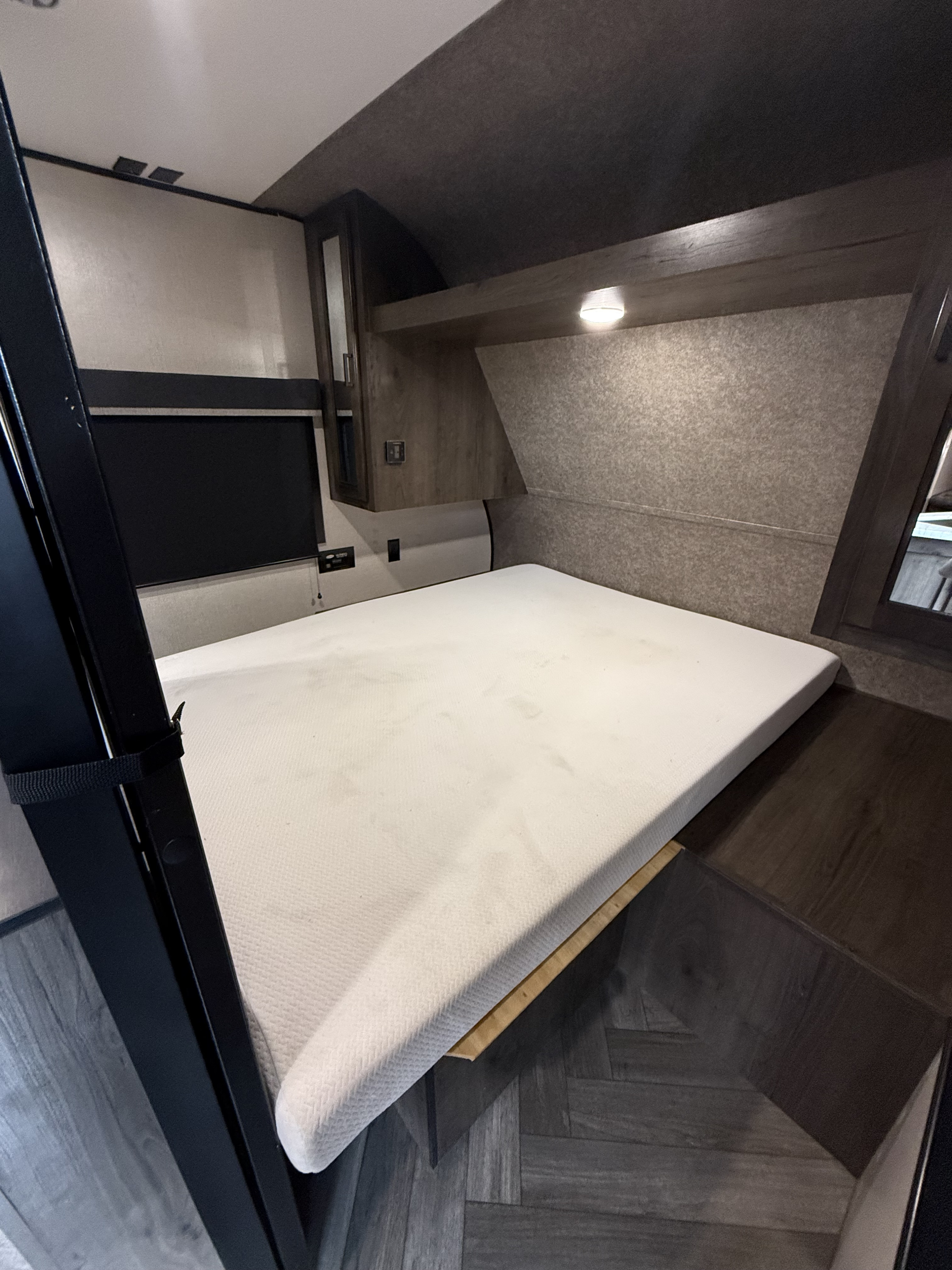 RV rental bedroom interior with a white mattress (showing stains), overhead storage, and dark herringbone floor. A side window is seen.. Olympia 26BHS 2022