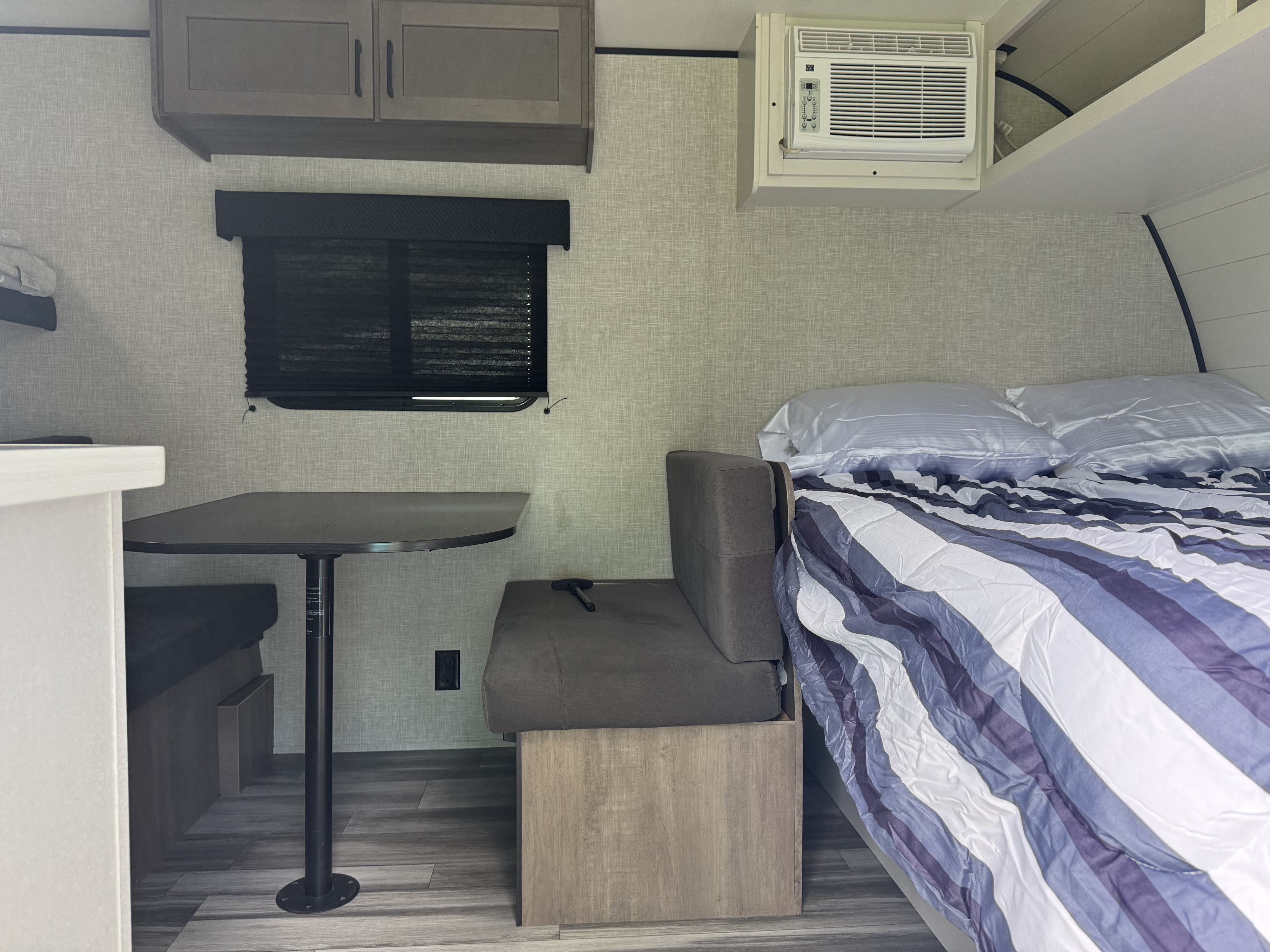 Experience this rv rental's compact interior: a striped bed, dinette with table, overhead cabinets, window, and AC unit.. Jayco Jay Flight SLX 2025