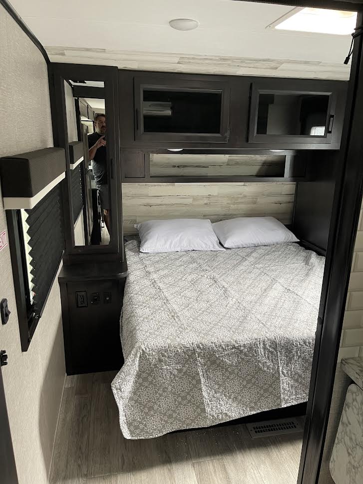 RV rental bedroom view: features a patterned bed, dark overhead cabinets, a light wood headboard, and a mirrored closet.. Jayco Jay Feather 2022