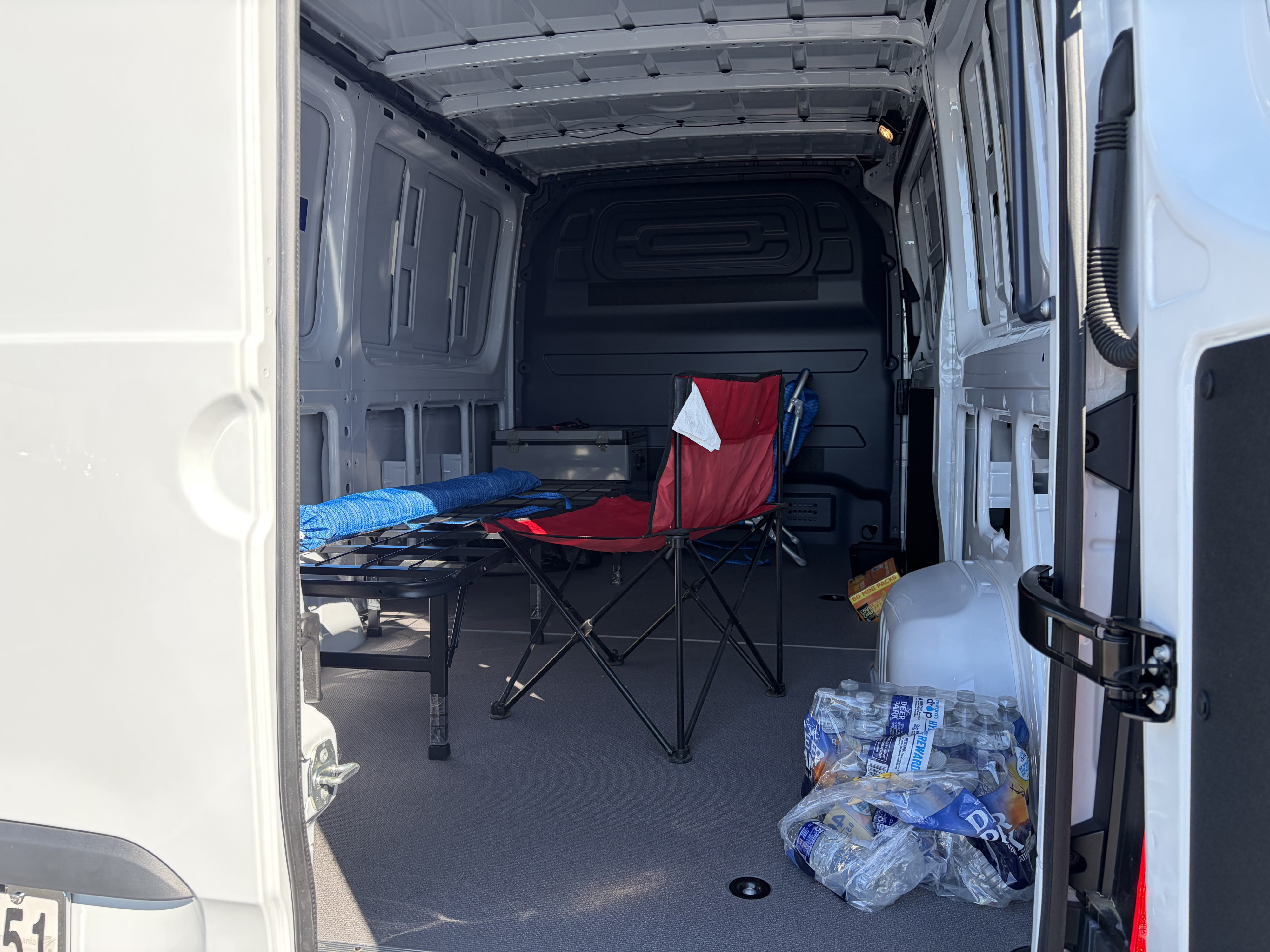 RV rental cargo van interior: cot, red chair, water bottles, and storage box on a carpeted floor.. Mercedes-Benz Sprinter 2025