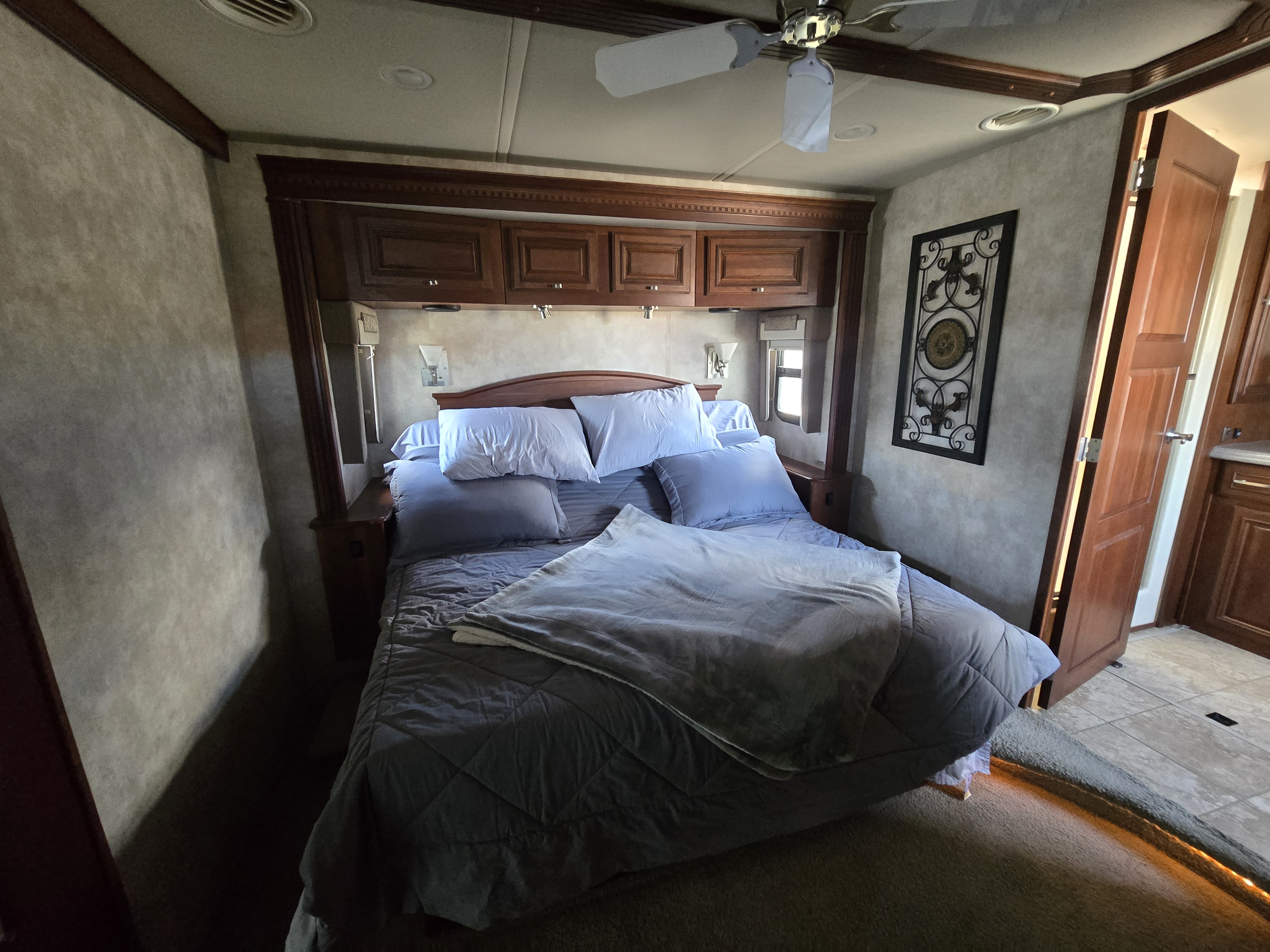 Cozy RV rental bedroom with a bed made with blue and white bedding, wooden overhead cabinets, a ceiling fan, and sconce lights.. Winnebago Tour 2011